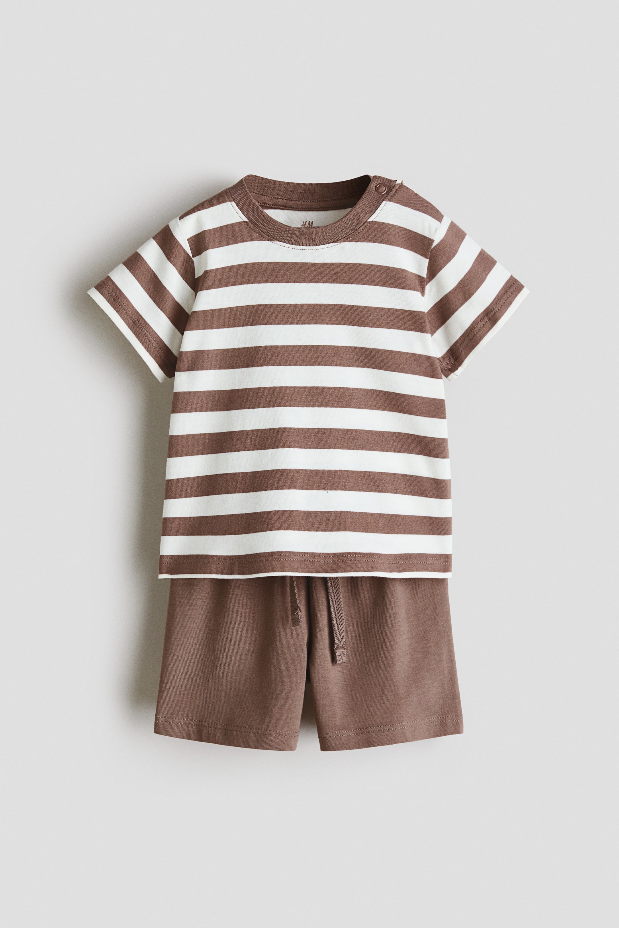 Größeres Bild anzeigen: Ein Set aus einem T-Shirt und Shorts aus weichem Baumwolljersey wird von vorne präsentiert. Das T-Shirt zeigt braune und weiße horizontale Streifen, einen gerippten Rundhalsausschnitt und Druckknöpfe auf der rechten Schulter, während die einfarbigen braunen Shorts einen elastischen Bund mit Kordelzug aufweisen.