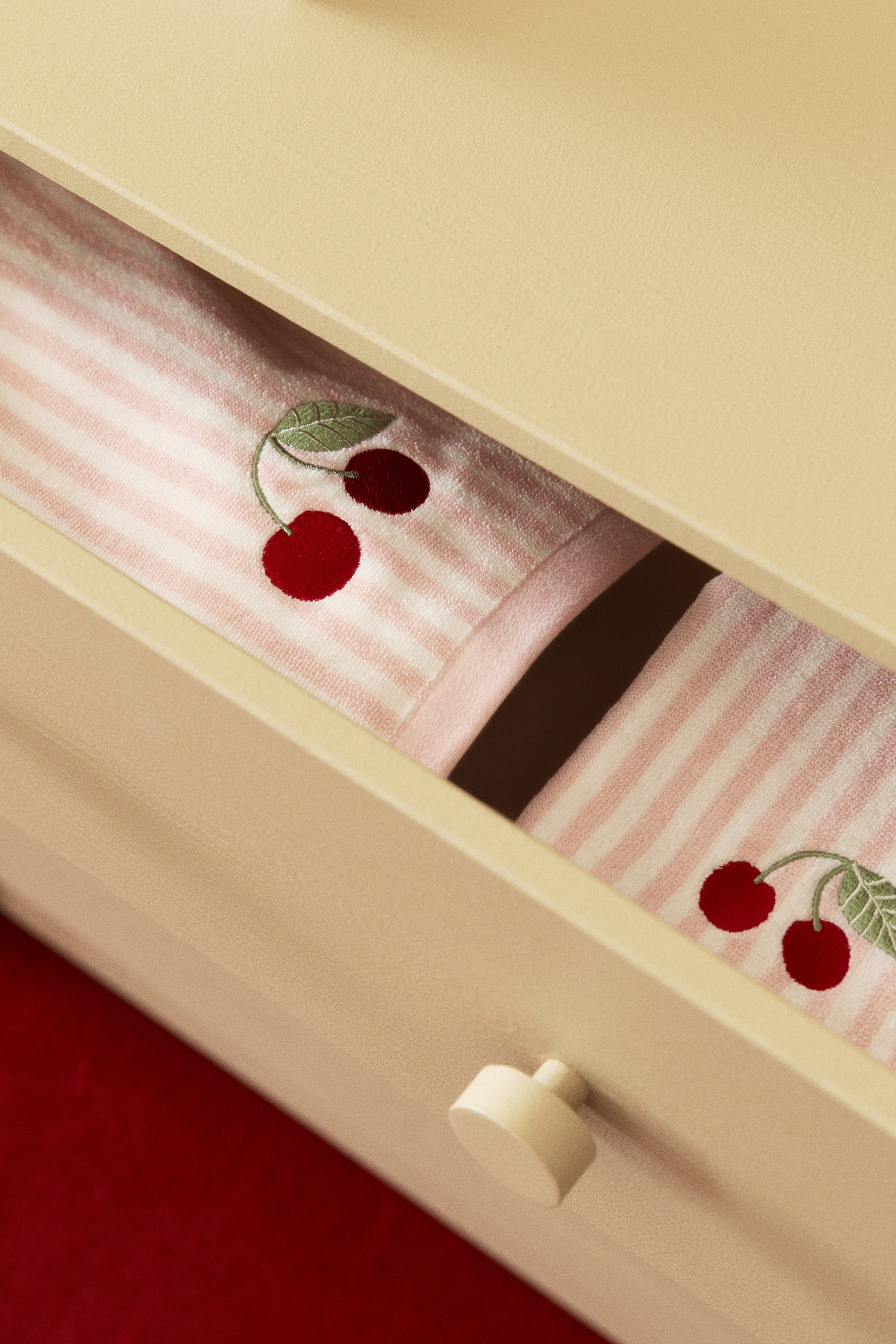 View larger image: A pink and white striped fabric, featuring red cherry embroidery with green stems and leaves, is lying folded and partially visible.