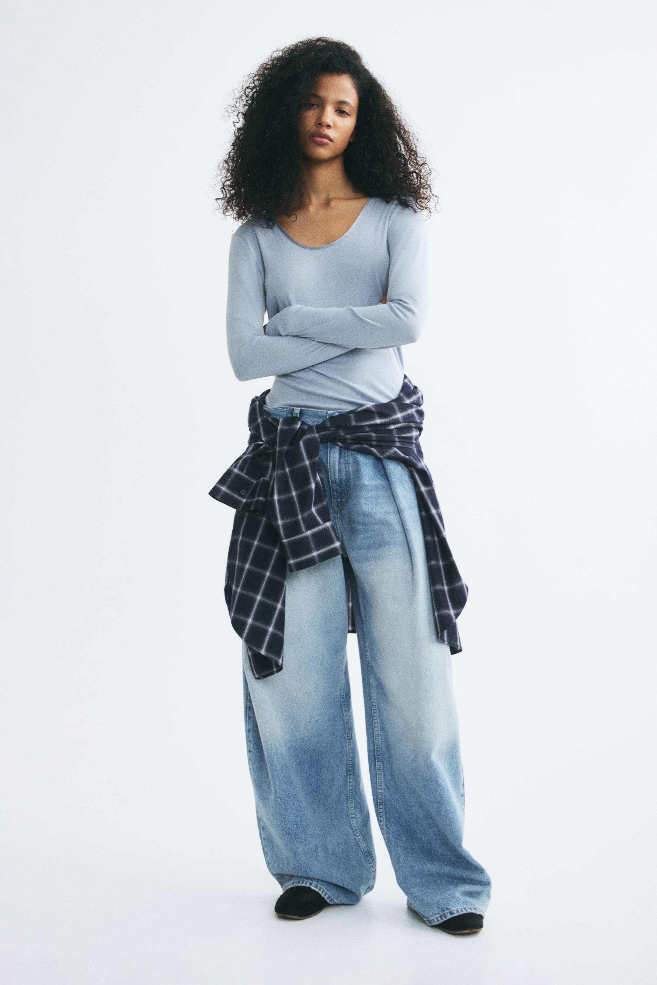 View larger image: A light blue fitted jersey top with a scoop neck and long sleeves is worn by the model, who is standing with crossed arms. The top is styled with wide-leg denim jeans and a dark plaid shirt tied around the waist.