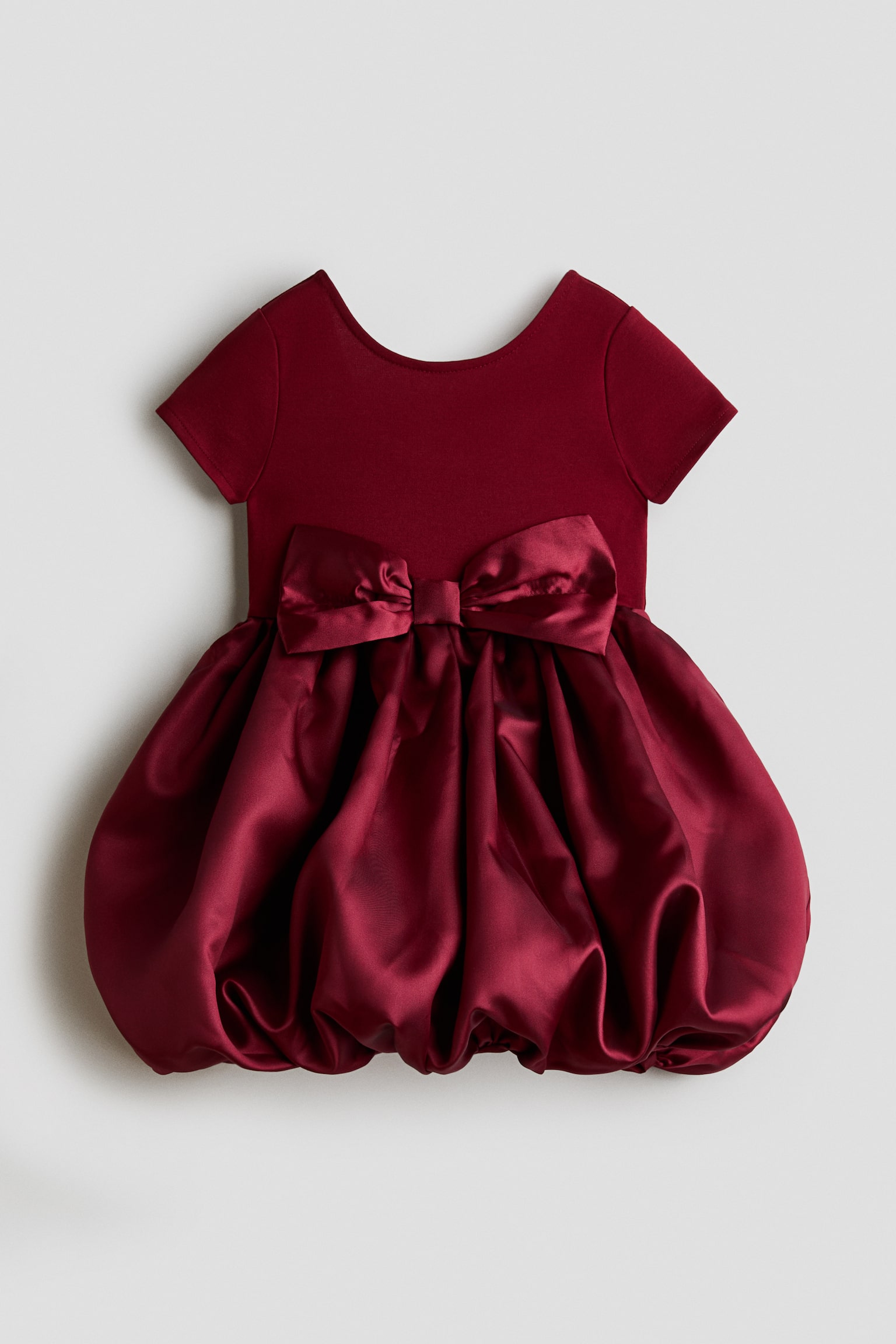 A burgundy dress is shown facing front, featuring a short-sleeved matte bodice with a scoop neckline and a voluminous satin bubble skirt. A large, matching satin bow sits at the centre front waistline, accentuating the transition from the knit top to the gathered, shiny skirt.