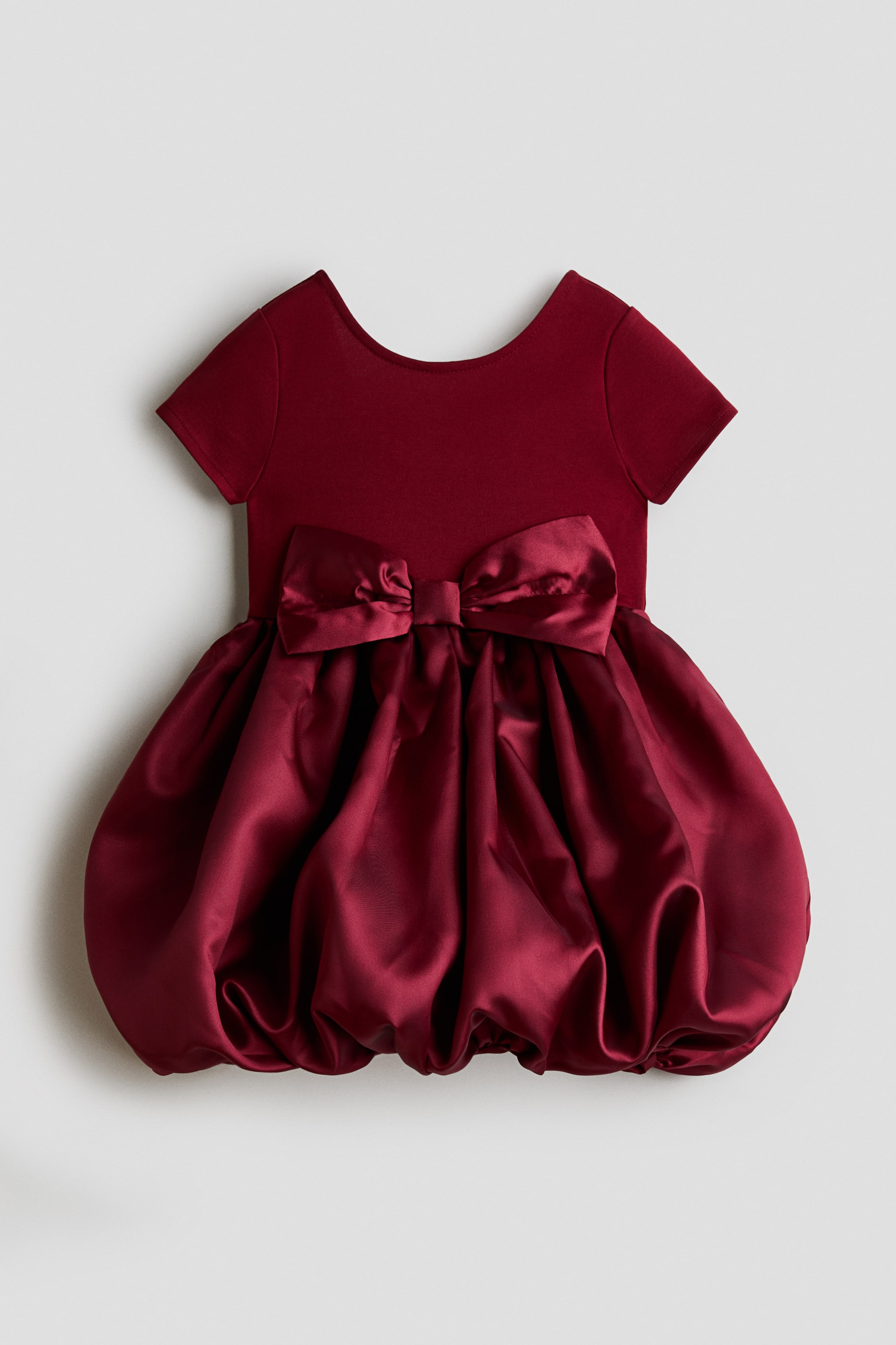 View larger image: A burgundy dress is shown front-facing, featuring a short-sleeved matte bodice with a scoop neckline and a voluminous satin bubble skirt. A large, matching satin bow sits at the centre front waistline, accentuating the transition from the knit top to the gathered, shiny skirt.