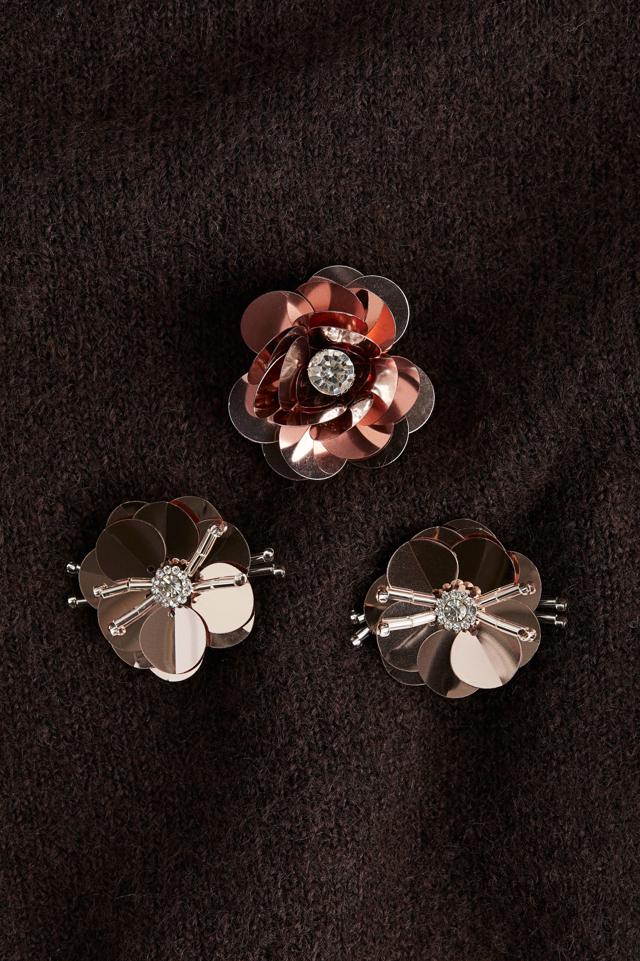 View larger image: Three rose gold-toned metallic flower-shaped embellishments are displayed on a dark brown knitted fabric. Each embellishment is featuring a central clear faceted gemstone; the two smaller ones are also exhibiting radiating clear bead sticks.