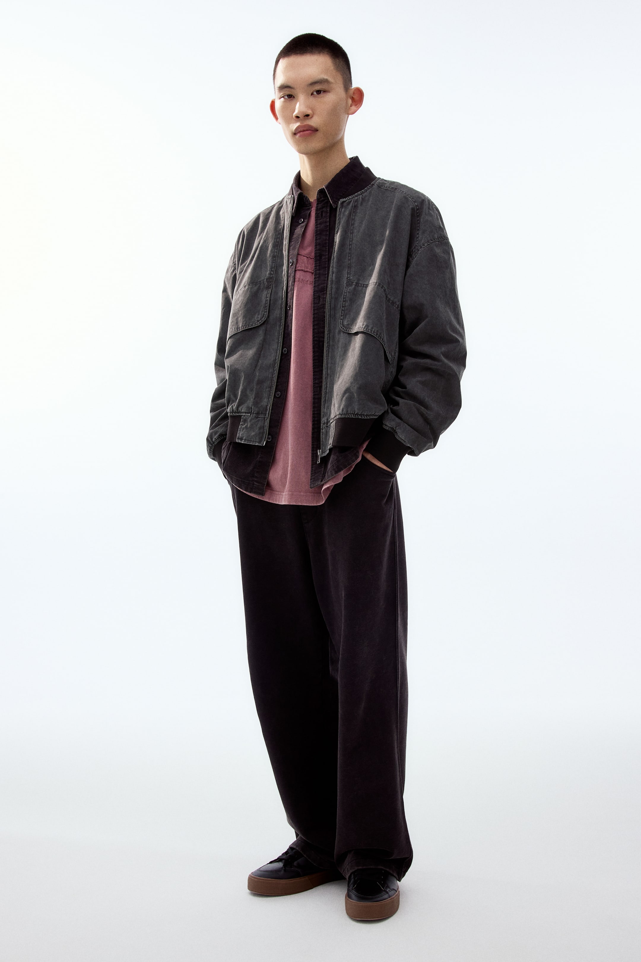 拡大画像を表示: モデルはウォッシュ加工の風合いがあるダークグレーのボンバージャケットを着用しています。リブ仕様のスタンドカラー、フロントのフラップポケット、袖口と裾はリブ仕上げです。ジャケットはジッパーを開け、赤みがかったピンクのシャツの上に重ねて着ています。