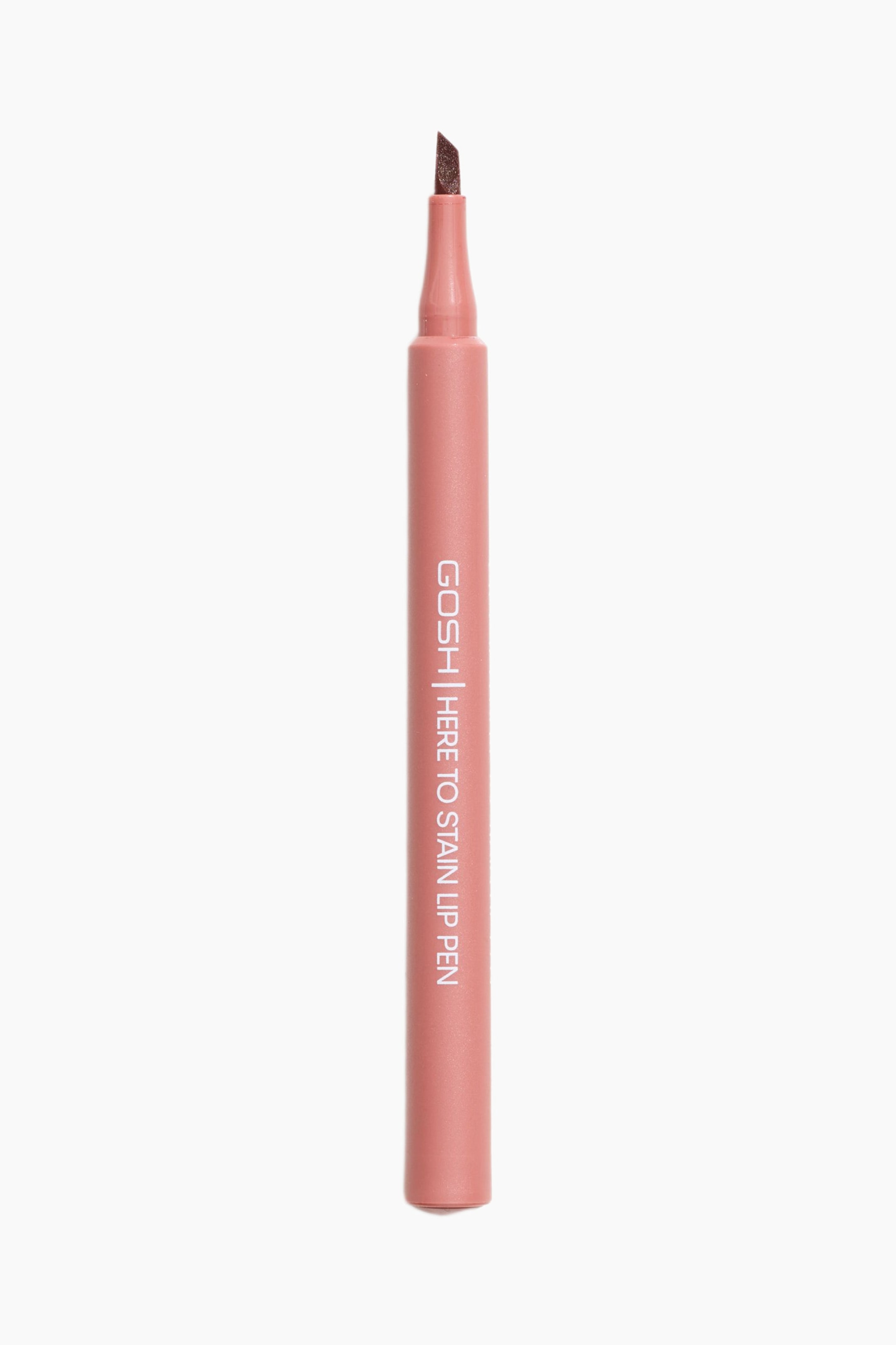 Visa större bild: En GOSH Here To Stain Lip Pen står upprätt, vänd framåt, och visar upp sin dämpade rosa cylindriska kropp med "GOSH | HERE TO STAIN LIP PEN" tryckt vertikalt. En mörkbrun, vinklad filtspetsapplikator är synlig på toppen.