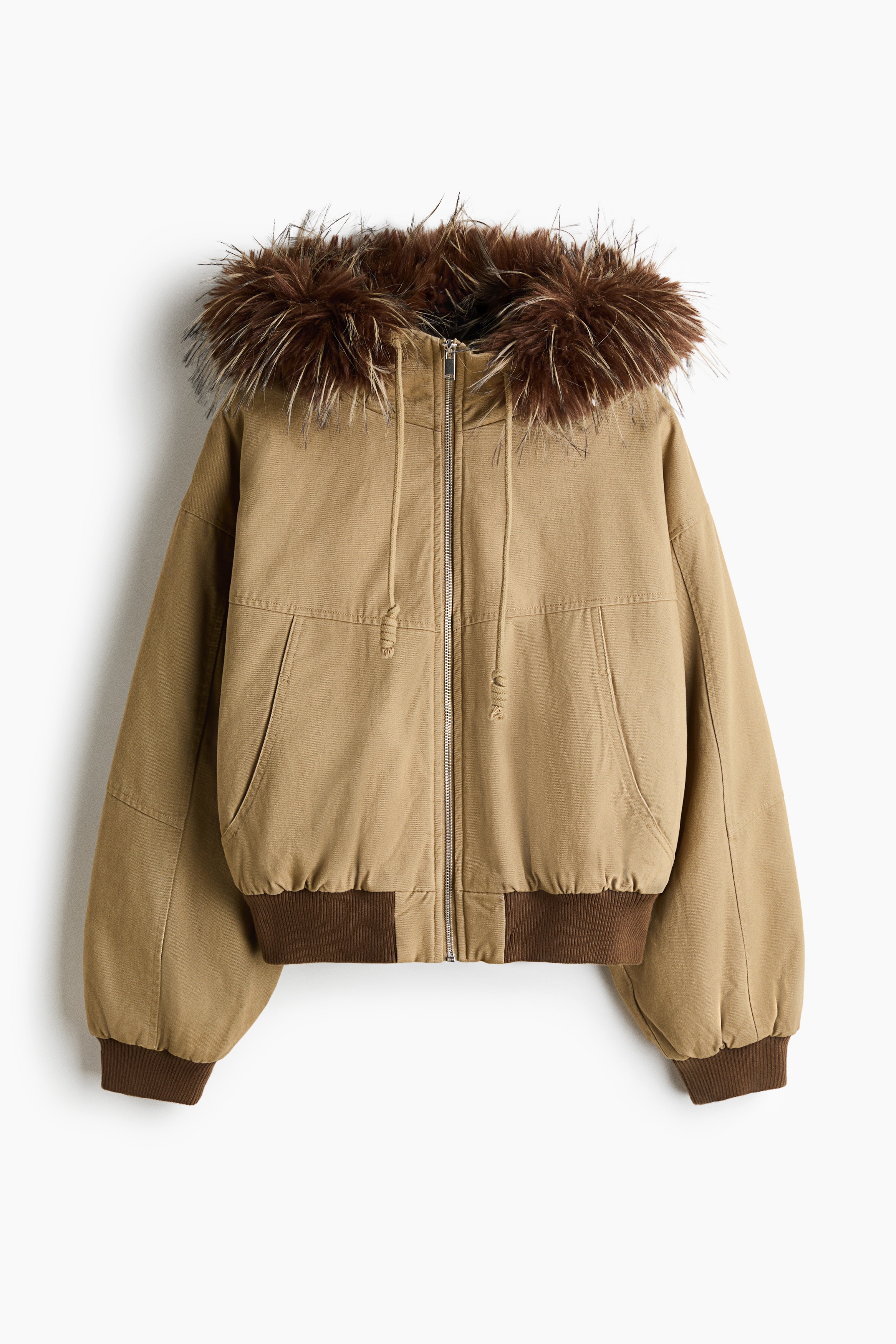 Model mÃ¡ na sebe bÃ©Å¾ovÃº oversized nylonovÃº bundu s rebrovanÃ½mi manÅ¾etami a veÄ¾kou kapucÅou s dlhÃ½m hnedÃ½m lemom z umelej koÅ¾uÅ¡iny. Model drÅ¾Ã­ a oboma rukami si upravuje nadÃ½chanÃº kapucÅu cez oÄi.