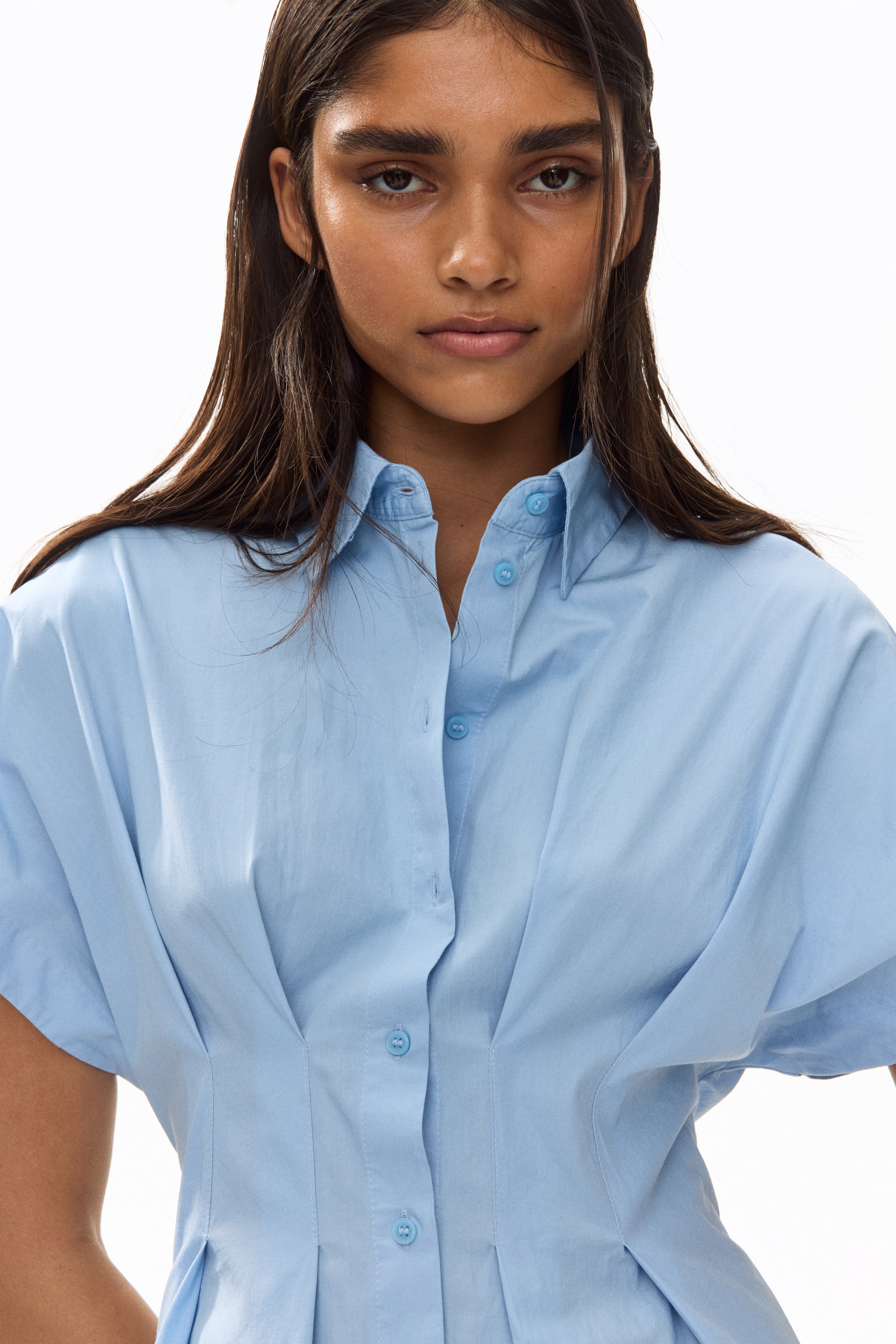 View larger image: The model is donning a light blue short-sleeved shirt dress crafted from a cotton weave. It boasts a collar, buttons running down the front, and inverted box pleats at the waist.
