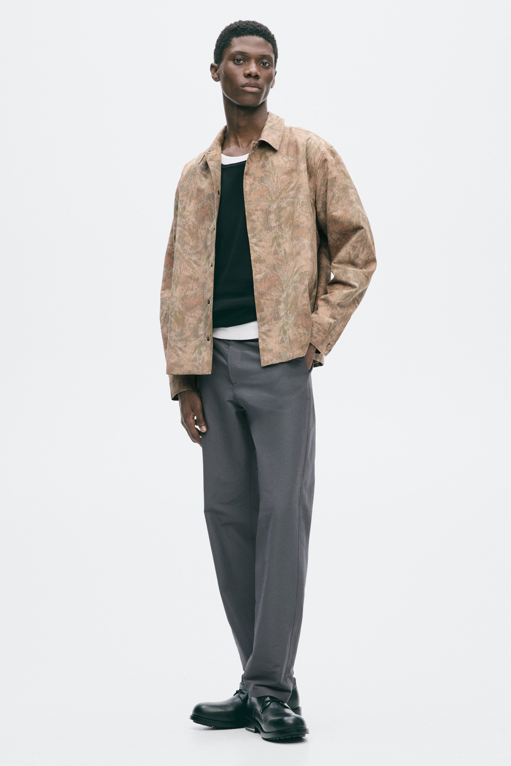 Grösseres Bild ansehen: Ein Overshirt aus gewebtem Stoff mit dezentem Muster wird vom Model getragen und über anderen Kleidungsstücken kombiniert. Es verfügt über einen Kragen, eine vordere Knopfleiste mit sichtbarem oberstem Knopf, geknöpfte Ärmelabschlüsse und einen gerade geschnittenen Saum, und das Model stützt eine Hand in einer Nahttasche.