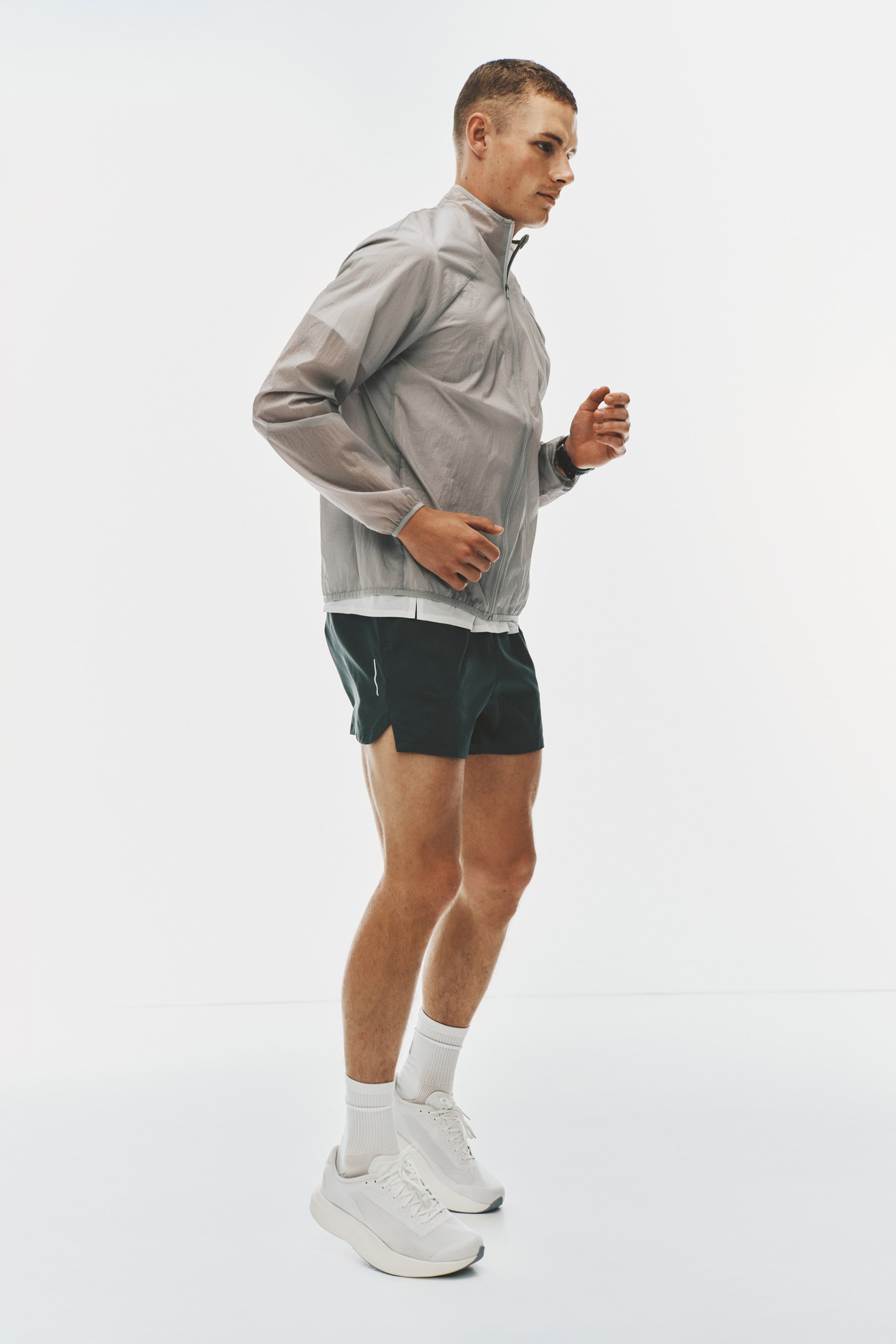 Ver imagen más grande: Una chaqueta de running translúcida de color gris claro con cremallera completa se lleva sobre una camiseta blanca, combinada con pantalones cortos deportivos de color gris oscuro que presentan una franja lateral blanca. Calcetines blancos hasta la mitad de la pantorrilla y zapatillas de running blancas con suelas gruesas completan el conjunto activo, que se presenta desde un ángulo lateral.