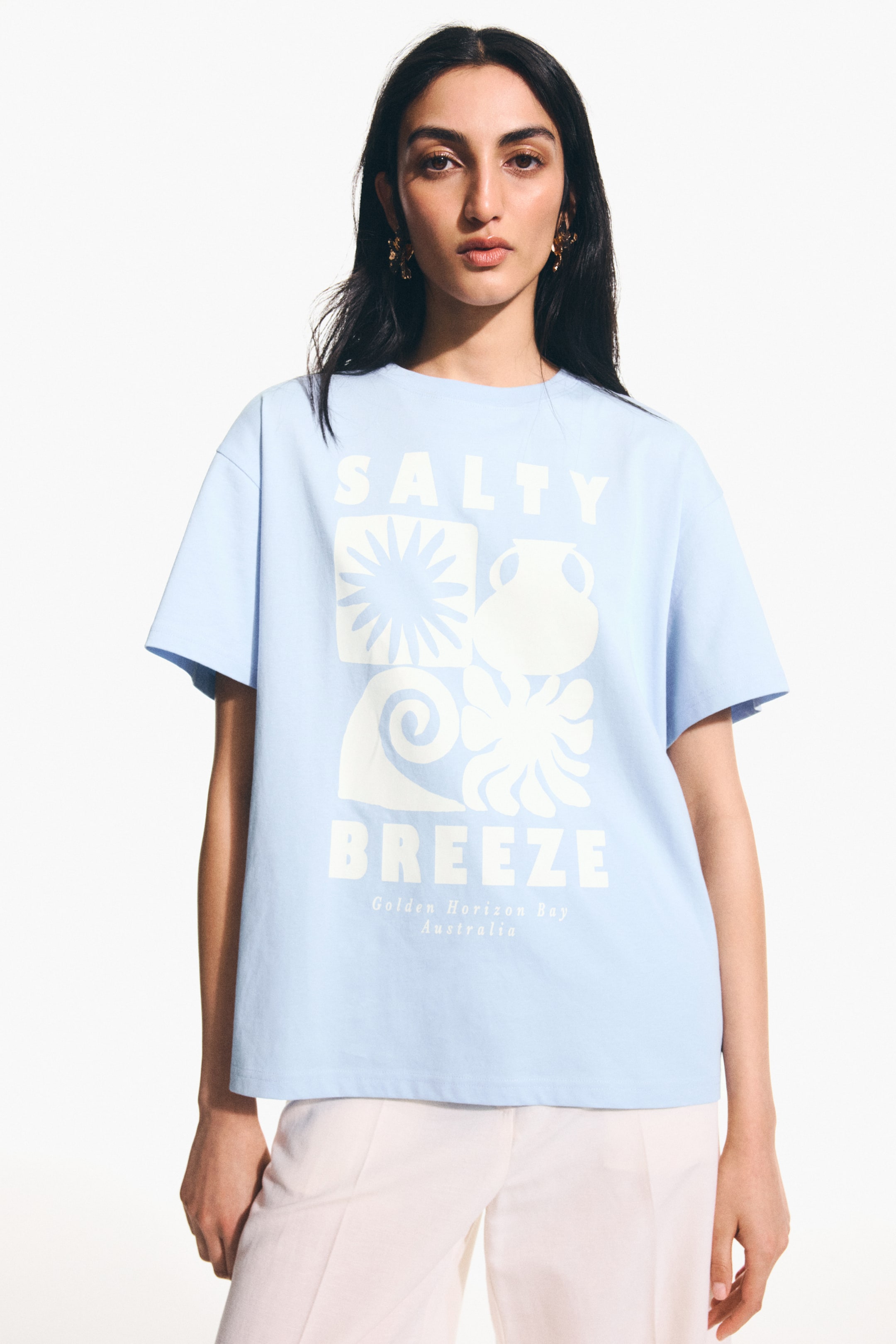 Ampliar la imagen: Una camiseta azul claro de corte holgado en jersey de algodón estampado, con un diseño gráfico blanco que incluye el texto "Salty Breeze" y formas abstractas, está siendo usada por la modelo. Cuenta con un cuello acanalado, hombros caídos y un dobladillo de corte recto.