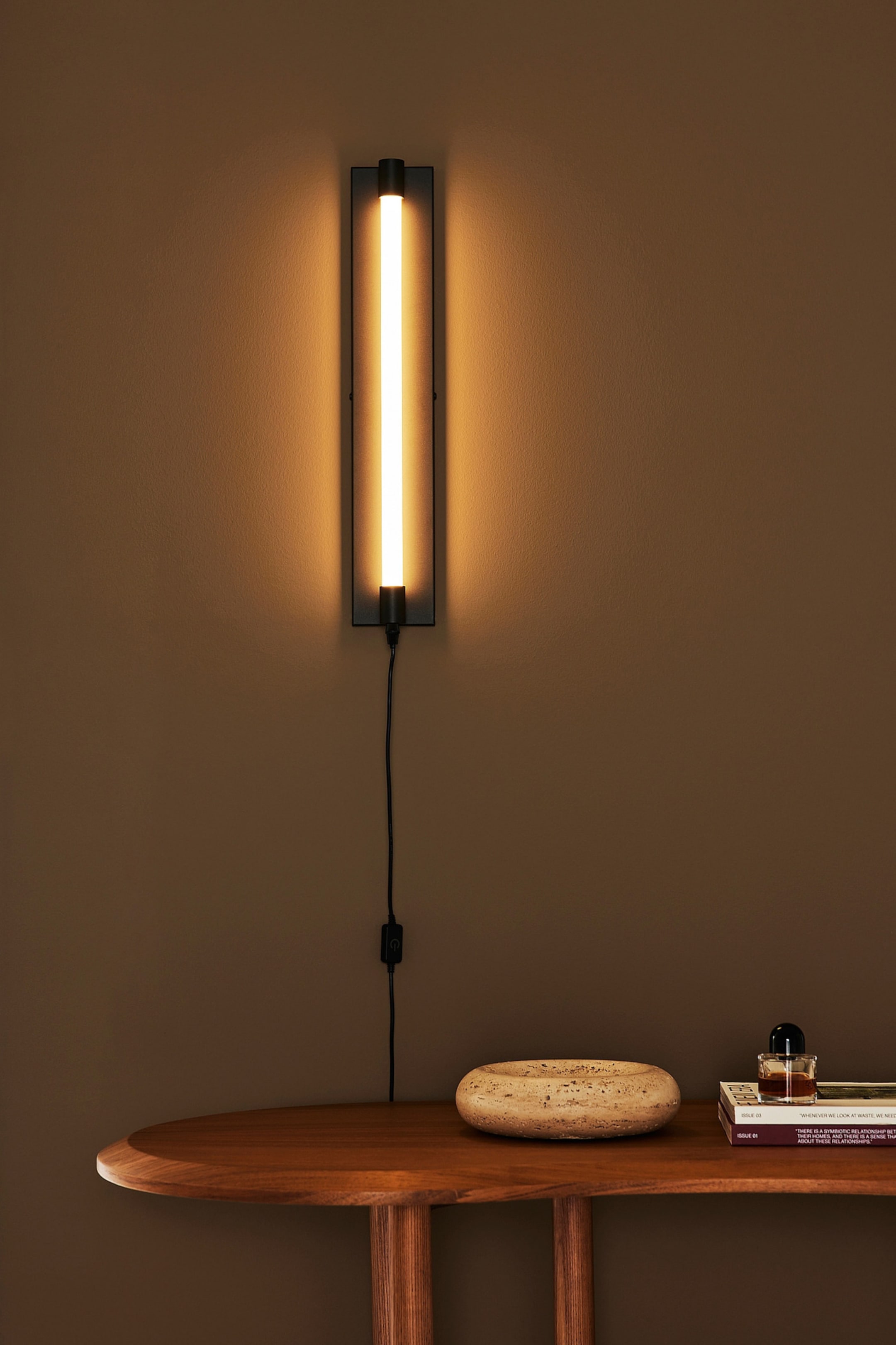 Se større billede: En vægmonteret lampe viser en lodret sort rektangulær base med en central glødende rørformet lyskilde, der udsender et varmt orangegult skær. En sort strømledning fører nedad fra bunden af armaturet, og en in-line-afbryder er synlig på ledningen.