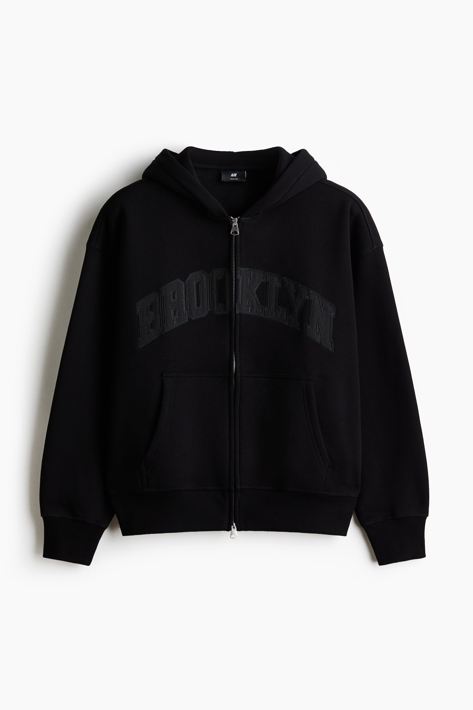 The black full-zip hoodie is shown facing front, boasting a silver-tone zip, a hood, and "BROOKLYN" appliquéd in matching black across the chest. It features ribbed cuffs, a ribbed hem, and two front patch pockets.