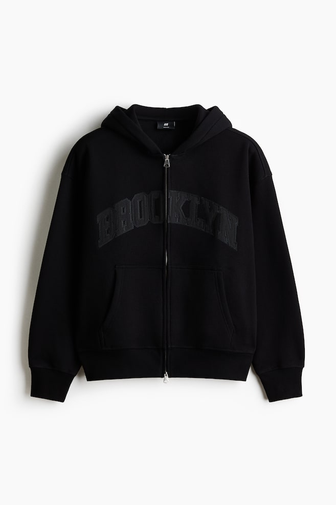 A black full-zip hoodie is facing front, featuring a silver-tone zipper, a hood, and "BROOKLYN" appliqued in matching black across the chest. It is displaying ribbed cuffs, a ribbed hem, and two front patch pockets. - 1