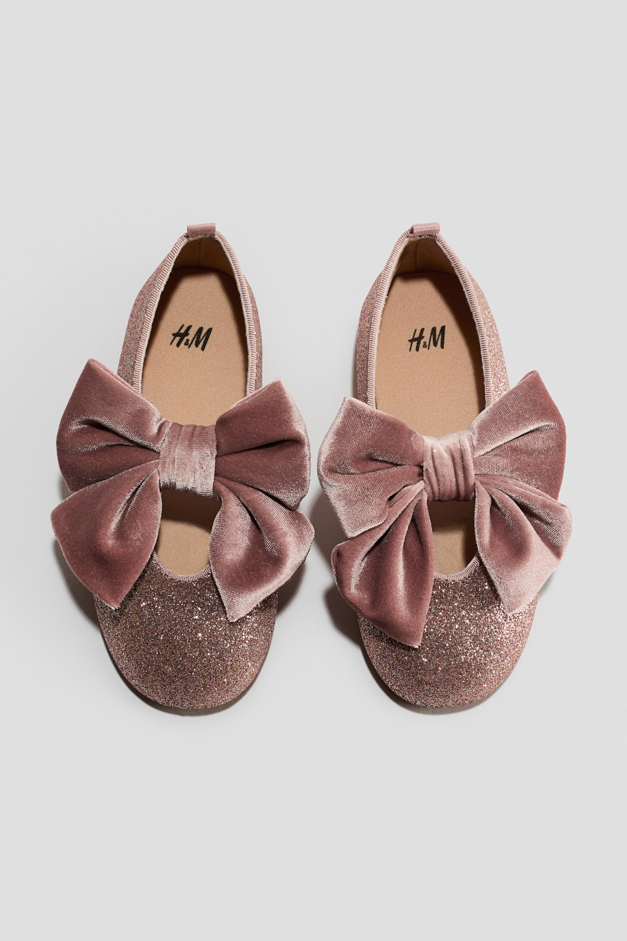 View larger image: A pair of pink glittery ballet pumps is resting side by side, showcasing a large pink velvet bow on each foot strap. The shoes feature a grosgrain trim around the top edge and a small loop at the back.