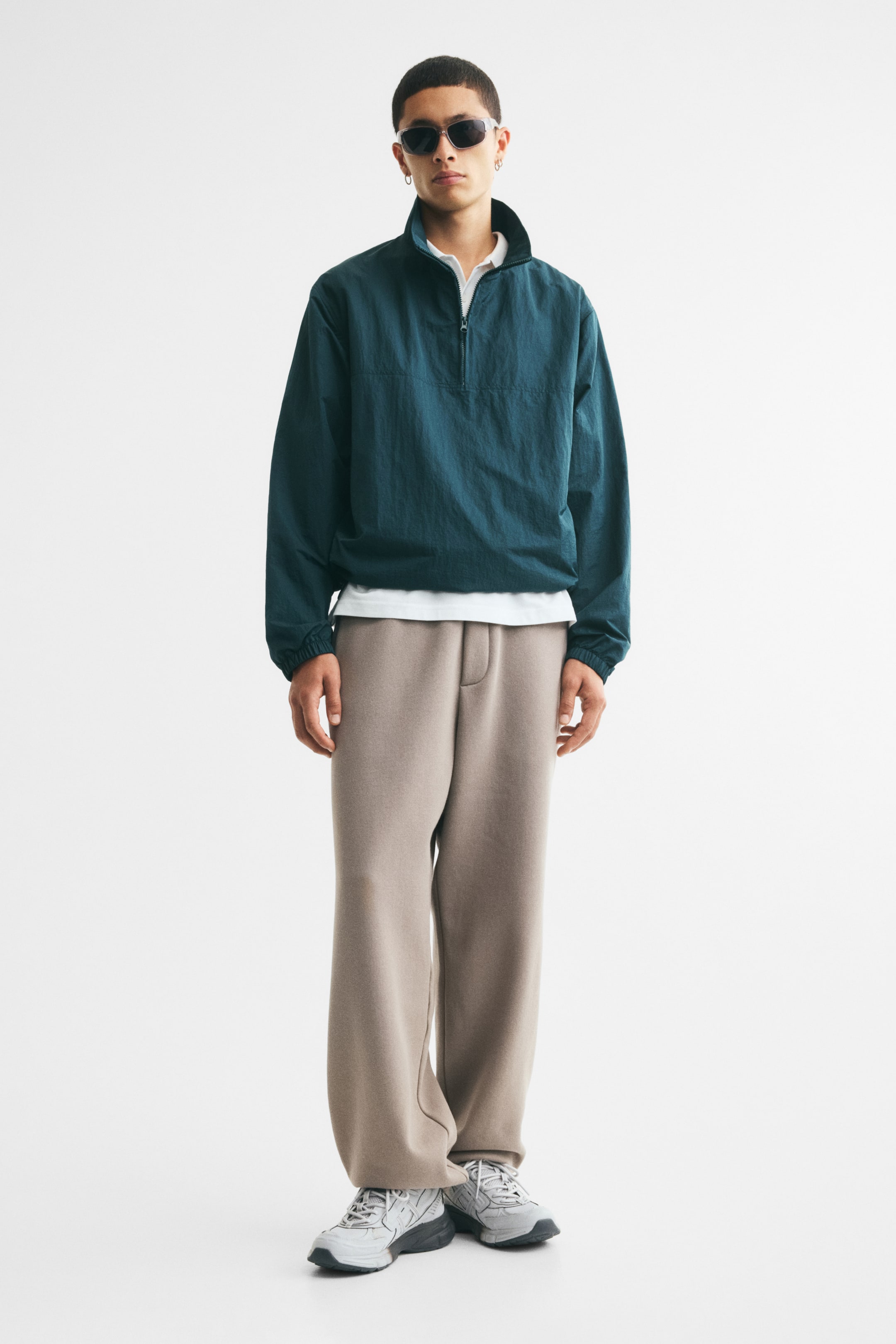Visualizza l'immagine ingrandita: Un windbreaker in nylon color verde petrolio scuro, con colletto alla coreana, chiusura mezza zip sul davanti e polsini elasticizzati, indossato dalla modella. Il windbreaker è abbinato a una camicia bianca con colletto ed è completato da pantaloni beige a gamba larga.