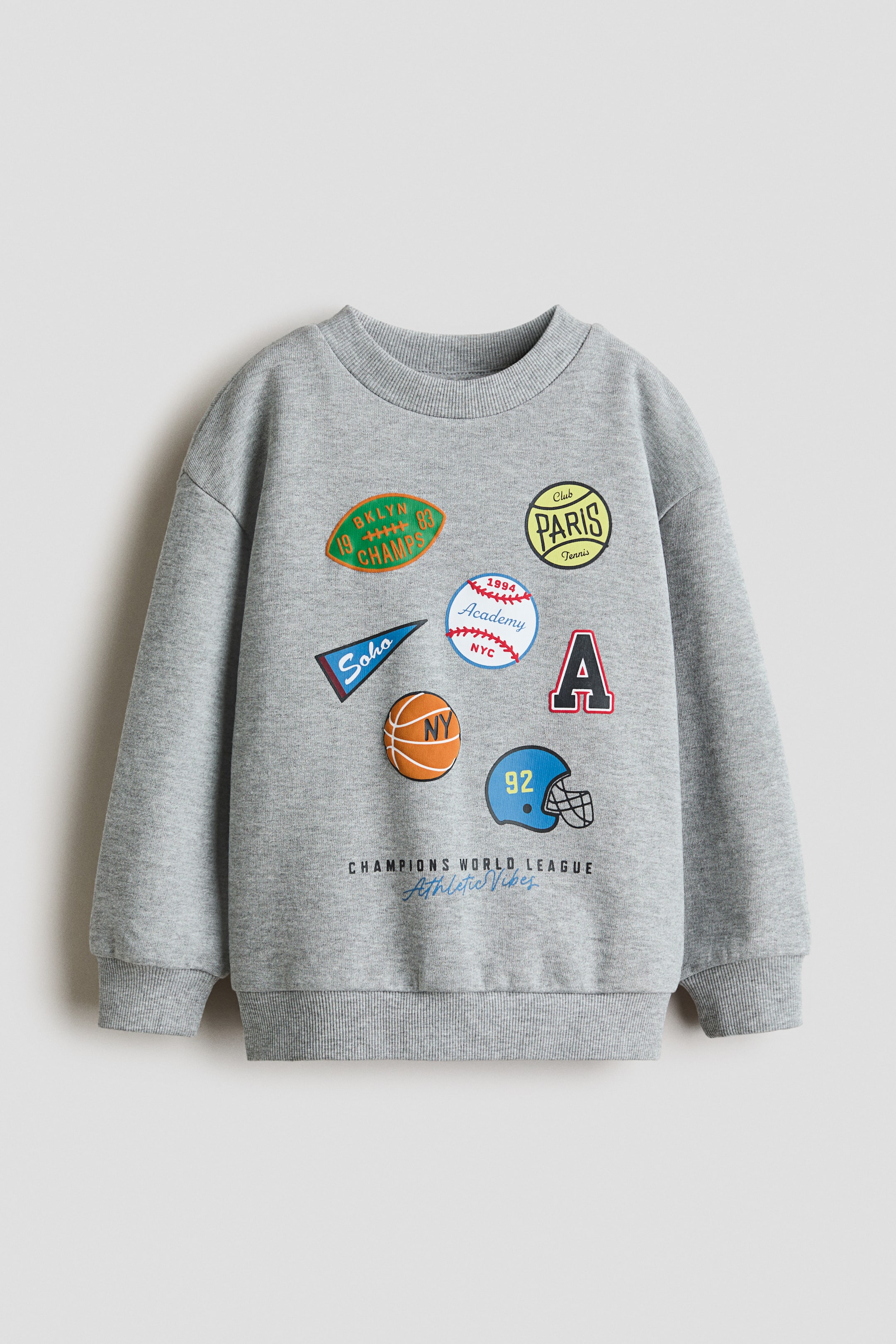 View larger image: A grey sweatshirt features a crew neck, long sleeves with ribbed cuffs, and a ribbed hem. The front showcases multiple colourful sports-themed appliqués and text, including a green "BKLYN CHAMPS" oval, a yellow "PARIS Tennis" circle, a red letter "A", an orange basketball with "NY", and a blue football helmet with "92", while "CHAMPIONS WORLD LEAGUE Athletic Vibes" also appears below the appliqués.