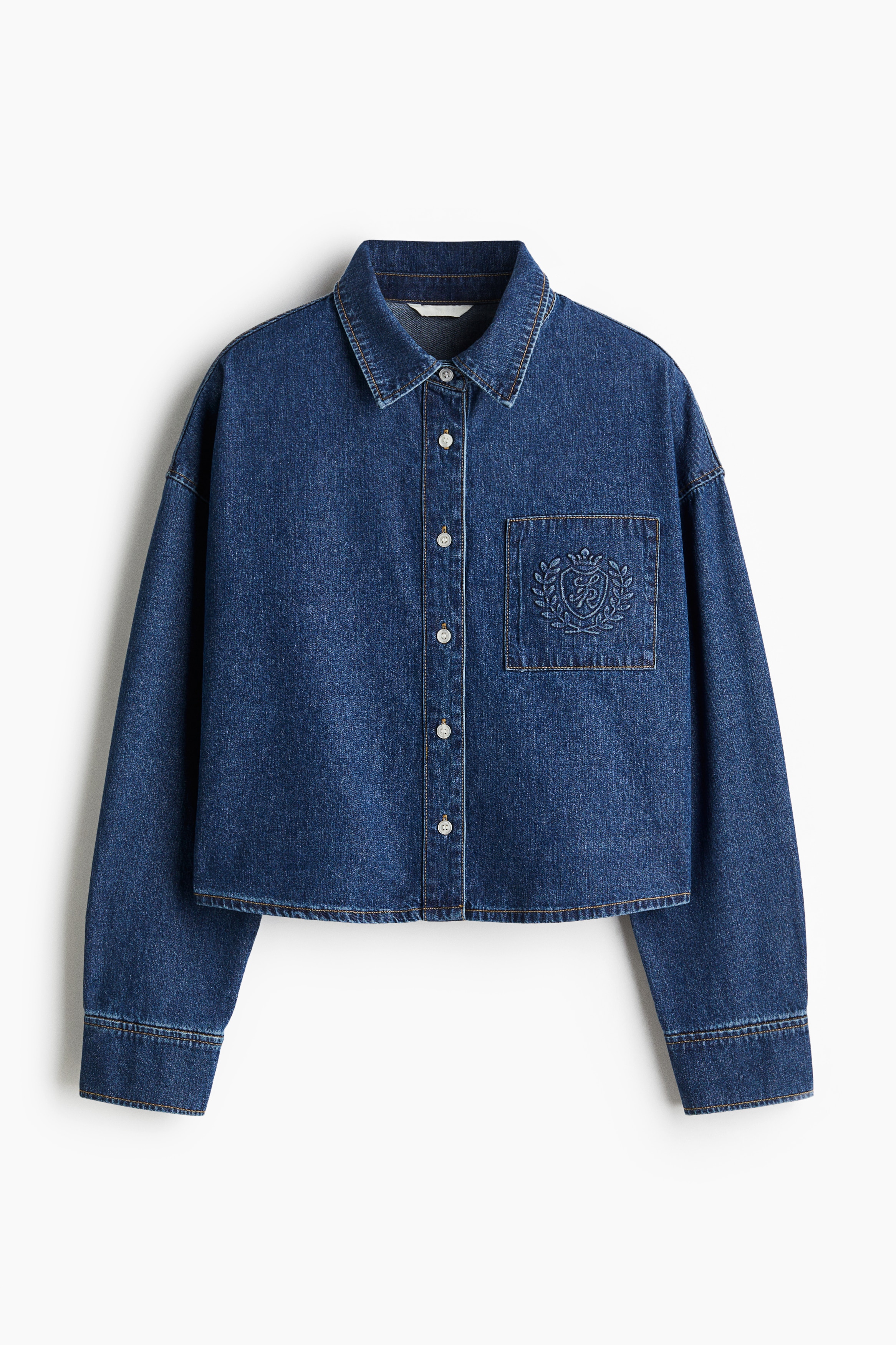 A blue denim shirt is being displayed from the front, angled slightly to the right. It is layered over a white t-shirt and is featuring a pointed collar, a button-down placket with white buttons, a patch pocket on the left chest with a tone-on-tone embroidered crest, and prominent topstitching along the seams.
