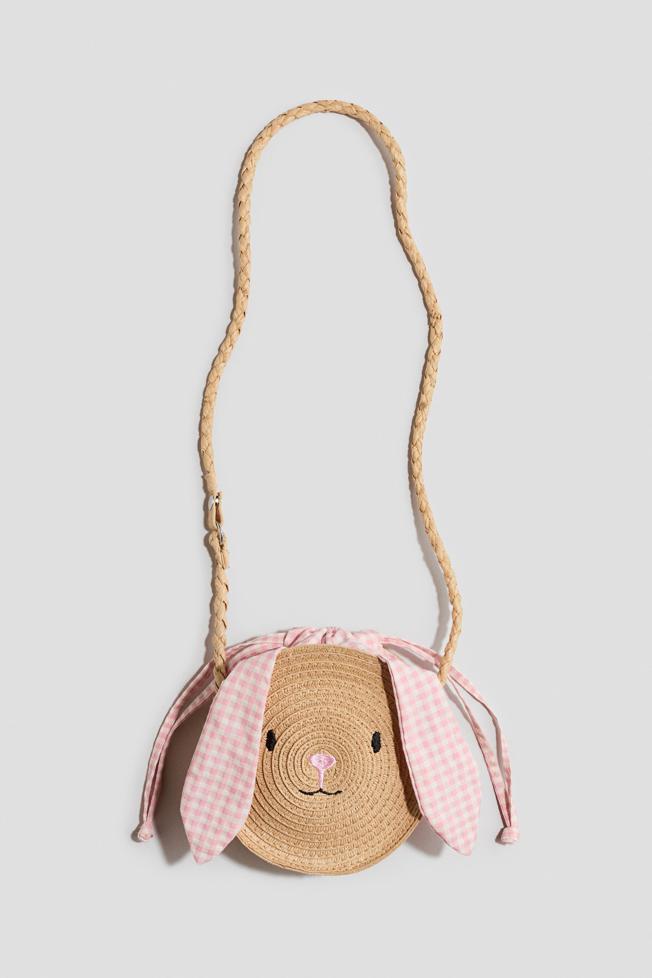 View larger image: A straw bunny-shaped crossbody bag is facing forward, featuring a round woven straw body with embroidered black eyes, a pink nose, and a black mouth. Pink and white gingham fabric ears are tied at the top, hanging loosely, and a braided straw strap is attached.