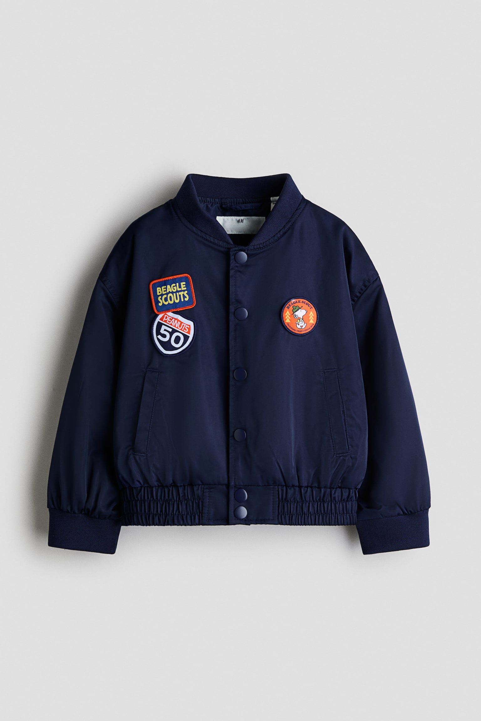 Má»t chiáº¿c Ã¡o khoÃ¡c bomber mÃ u xanh navy ÄÆ°á»£c chá»¥p á» gÃ³c chÃ­nh diá»n, ná»i báº­t vá»i cá» Ã¡o, cá» tay Ã¡o vÃ  gáº¥u Ã¡o bo gÃ¢n co giÃ£n. Ão cÃ³ hÃ ng cÃºc báº¥m á» giá»¯a thÃ¢n, tÃºi viá»n hai bÃªn, vÃ  ÄÆ°á»£c trang trÃ­ báº±ng cÃ¡c miáº¿ng patch chá»§ Äá» Peanuts á» cáº£ hai bÃªn ngá»±c.