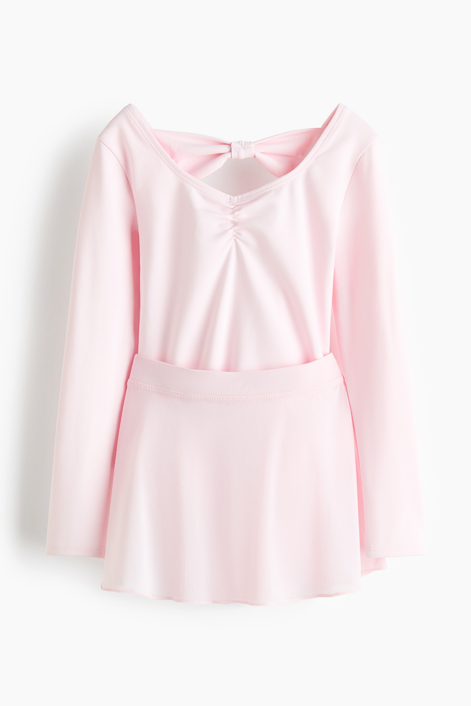 Un justaucorps de ballet rose à manches longues avec une jupe attachée présente son dos, mettant en valeur un décolleté en V avec un détail de nœud au centre du dos et des fronces douces se formant sous le nœud. La jupe attachée est légèrement fluide. - 1