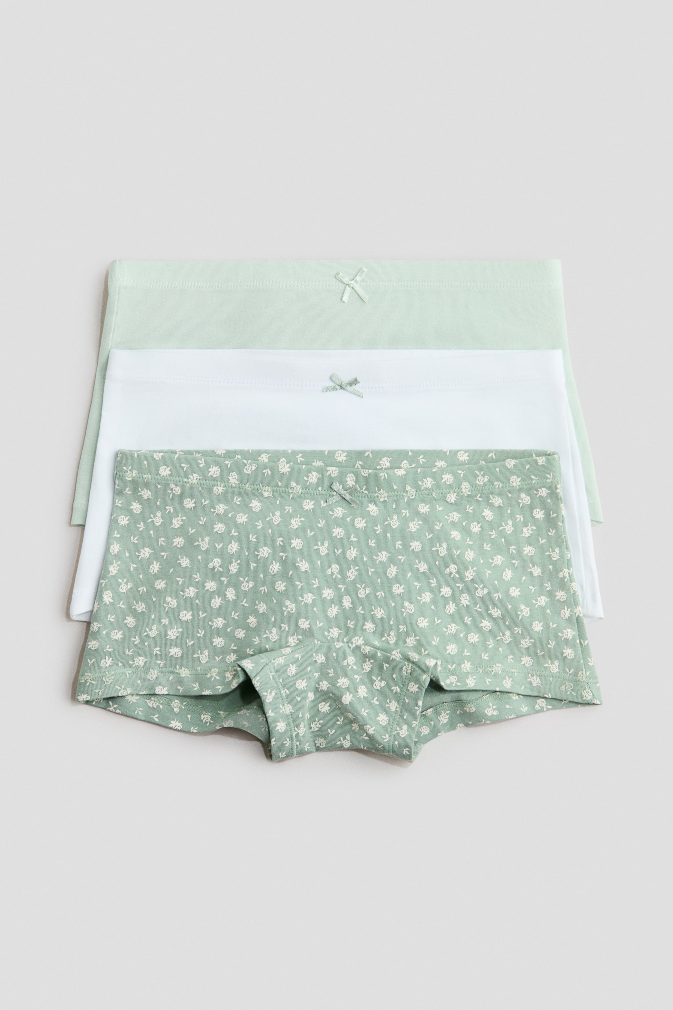 View larger image: Three pairs of cotton jersey boxer briefs are stacked, facing front. The top pair is light green with a white floral pattern, the middle pair is solid white, and the bottom pair is solid light green, with each pair featuring an elasticated waist and a small decorative bow at the centre front.