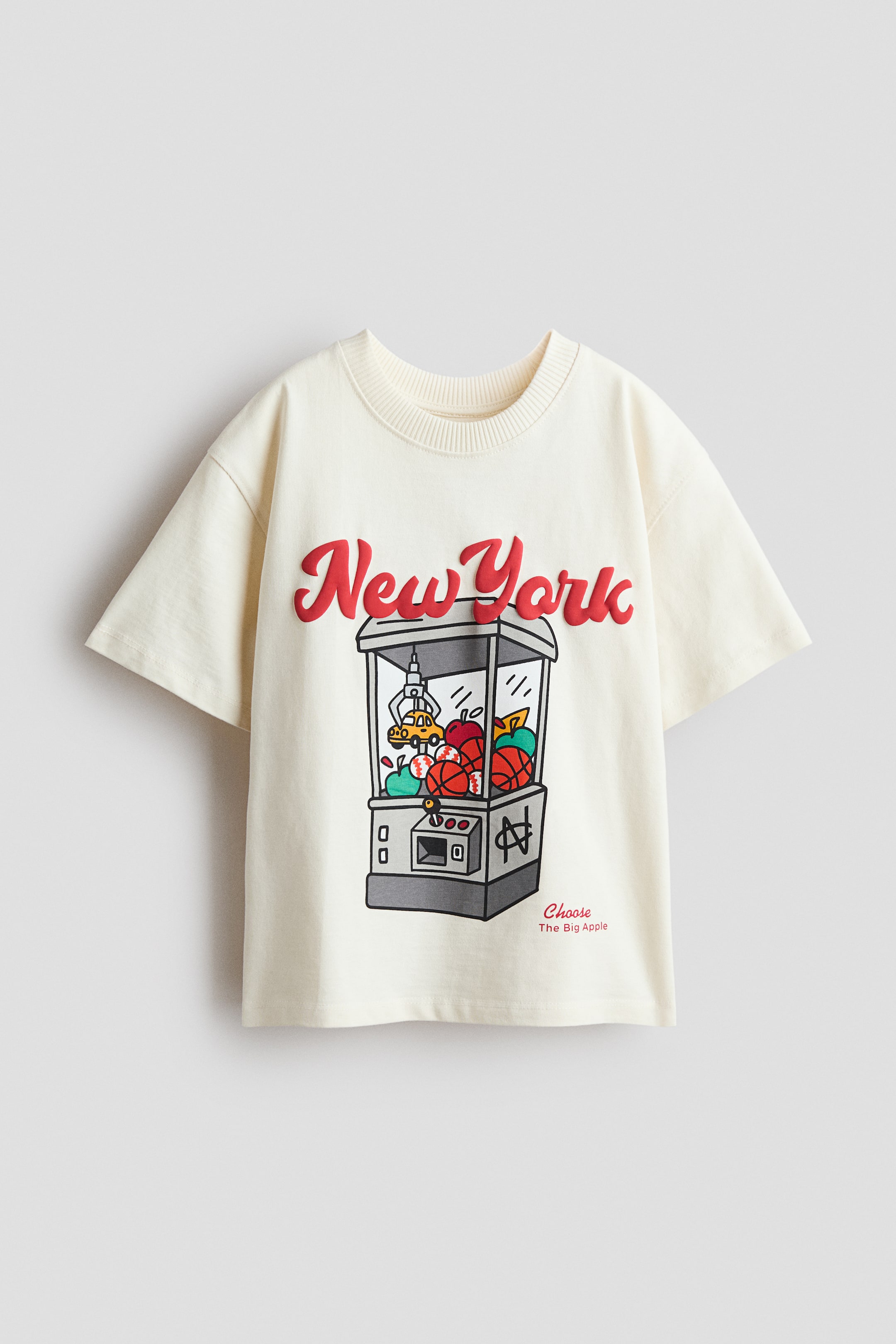 Grotere afbeelding bekijken: Een losvallend, crèmekleurig t-shirt toont de voorkant, met een ronde, geribbelde halslijn en verlaagde schouders. Het is bedrukt met "New York" in rode, reliëfletters, gepositioneerd boven een grijpmachinegrafiek met appels en basketbalballen.