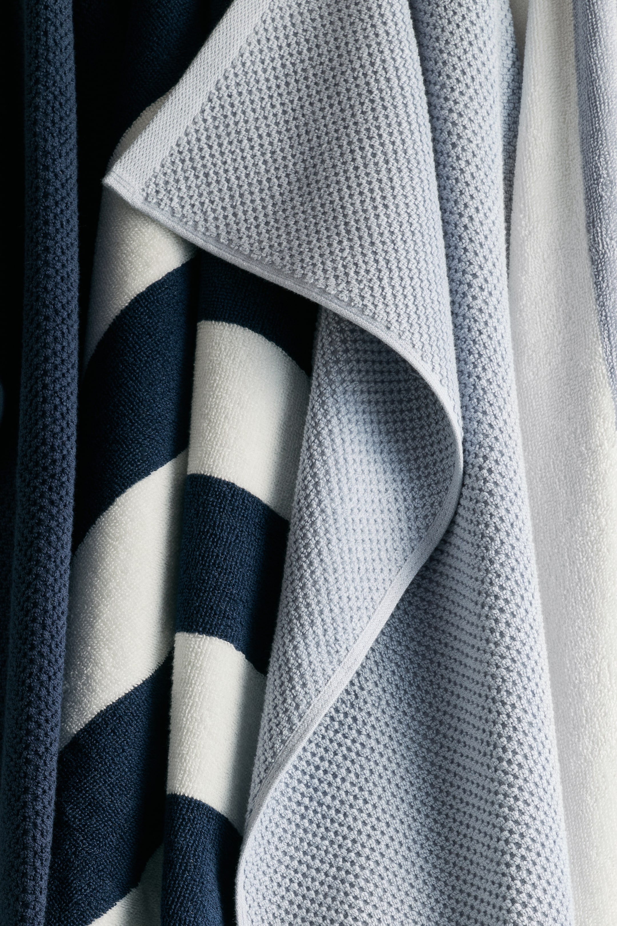 Prikaži veću sliku: Svetloplava-siva pamučna frotirana peškir elegantno se prekriva, prikazujući mali, ponavljajući tkan uzorak. Ovaj peškir je delimično preklopljen preko pamučnog frotiranog peškira sa mornarsko plavim i belim prugama, koji prikazuje svoju mekani, uvojni materijal, dok tamno plavi peškir sa teksturom vafla visi s leve strane.