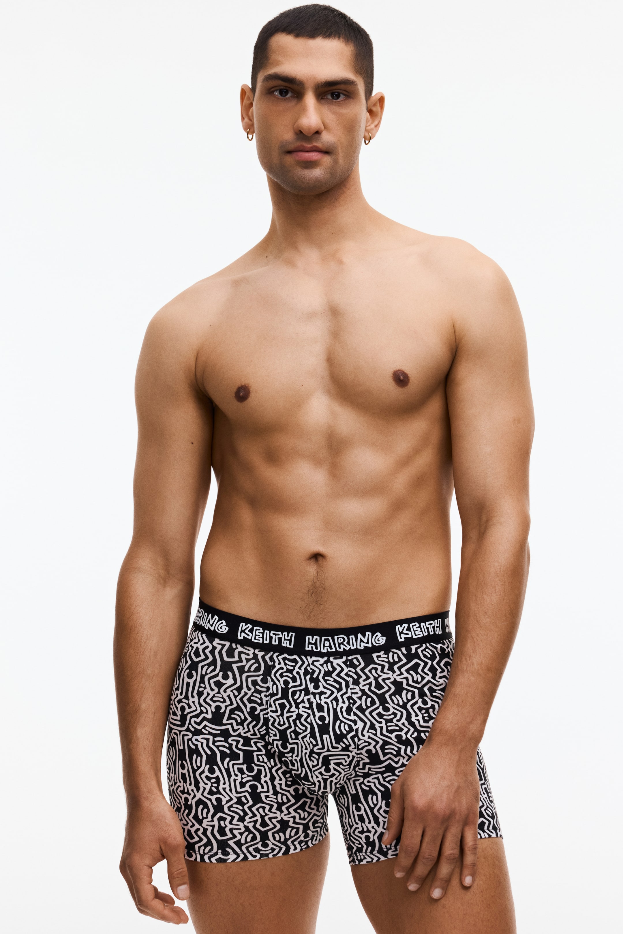 Büyük resmi görüntüle: Siyah boxer külotlar, stilize figürlerin beyaz desenleriyle kaplanmıştır. Siyah elastik bel bandı, "KEITH HARING" yazısını beyaz renkte tekrarlayarak sergiliyor ve boxer külotlar öne doğru bakarak vücuda oturan yapısını gösteriyor.
