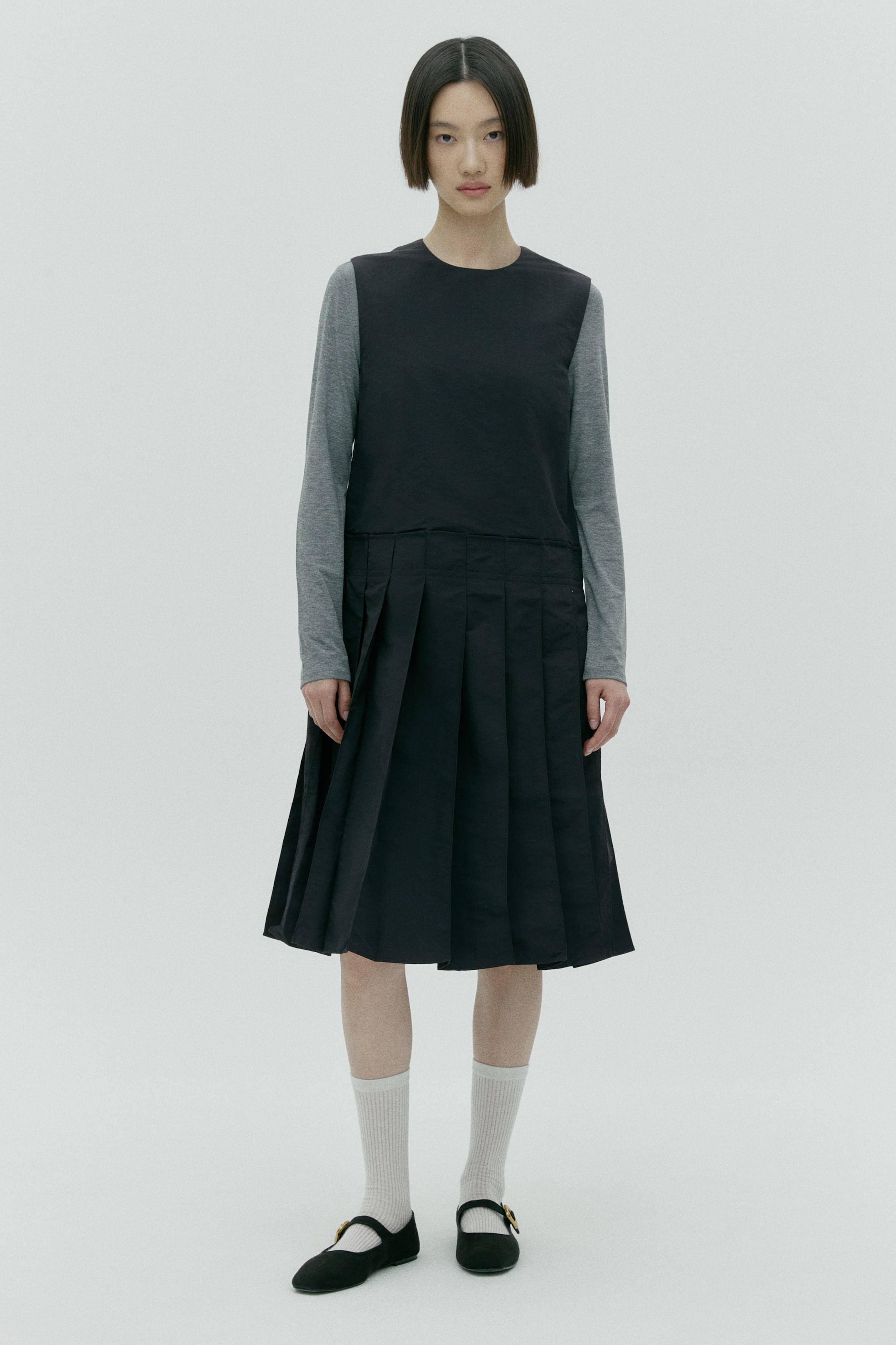 이미지 확대 보기: 블랙 민소매 드레스는 라운드 네크라인과 상체 중간에서 시작해 미디 길이까지 이어지는 플리츠 스커트 부분이 특징입니다. 회색 긴팔 셔츠 위에 레이어드되어 있습니다.