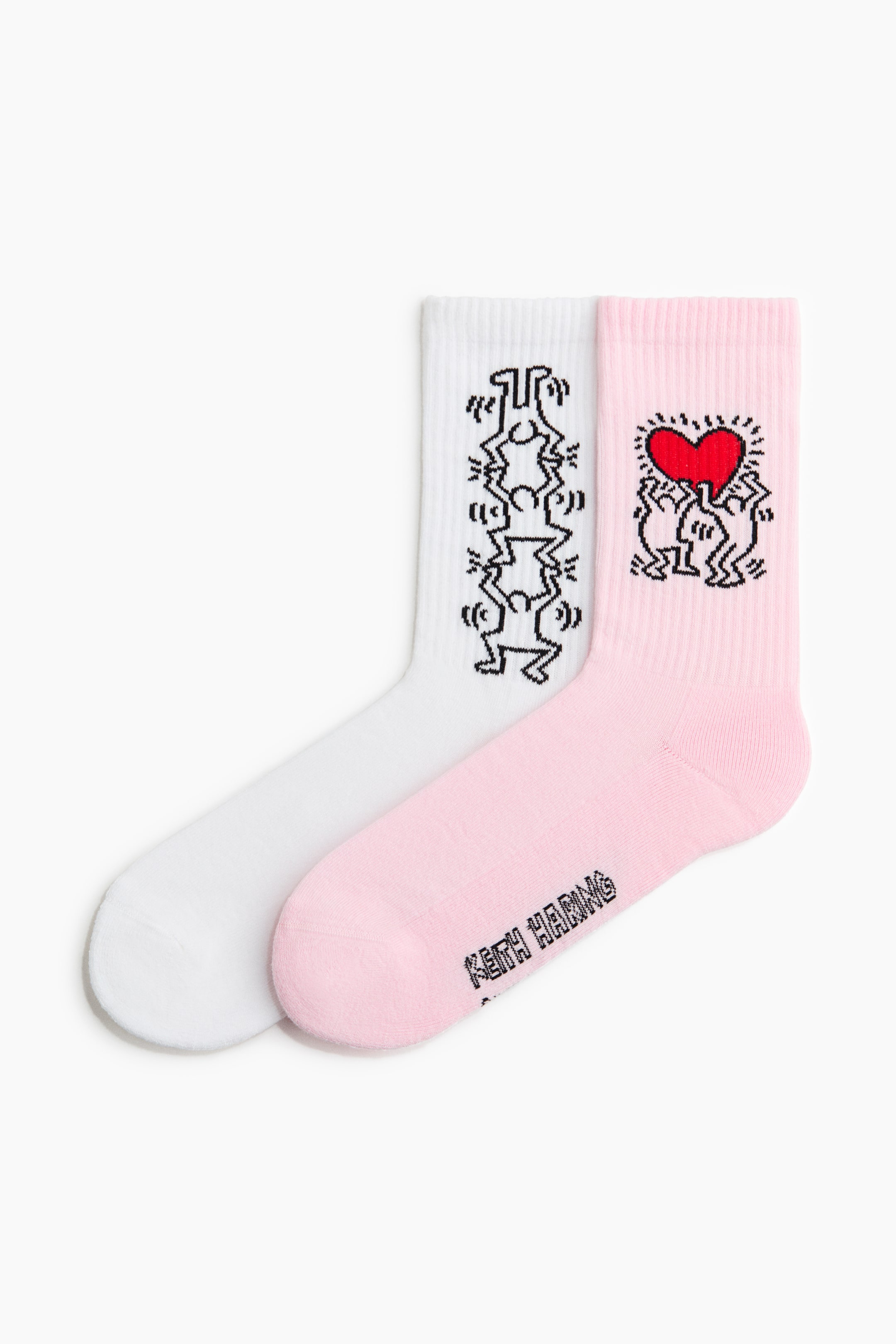 View larger image: A pair of socks is lying flat, one white and one light pink. The white sock boasts a black outline motif of stacked figures on its ribbed shaft, while the pink sock showcases a black outline motif of two figures holding a red heart on its ribbed shaft, with "KEITH HARING" text visible on its foot.