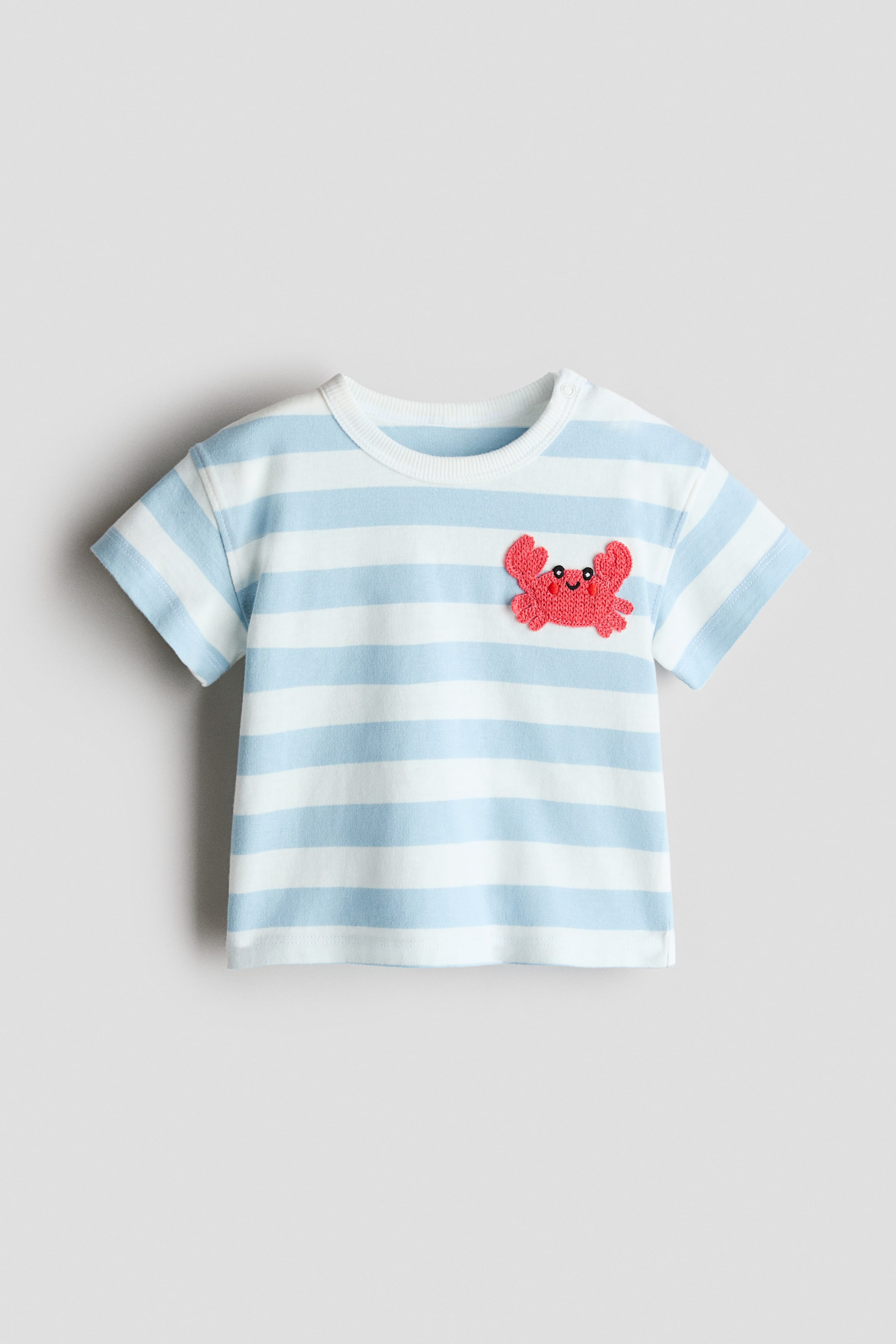 Ver imagen más grande: Una camiseta muestra su PARTE DELANTERA, con rayas horizontales en azul claro y blanco. Tiene un cuello redondo acanalado en blanco con cierre de botón a presión en el hombro izquierdo y mangas cortas. Un aplique de cangrejo rojo, texturizado, con ojos negros y una sonrisa está colocado en el pecho izquierdo.