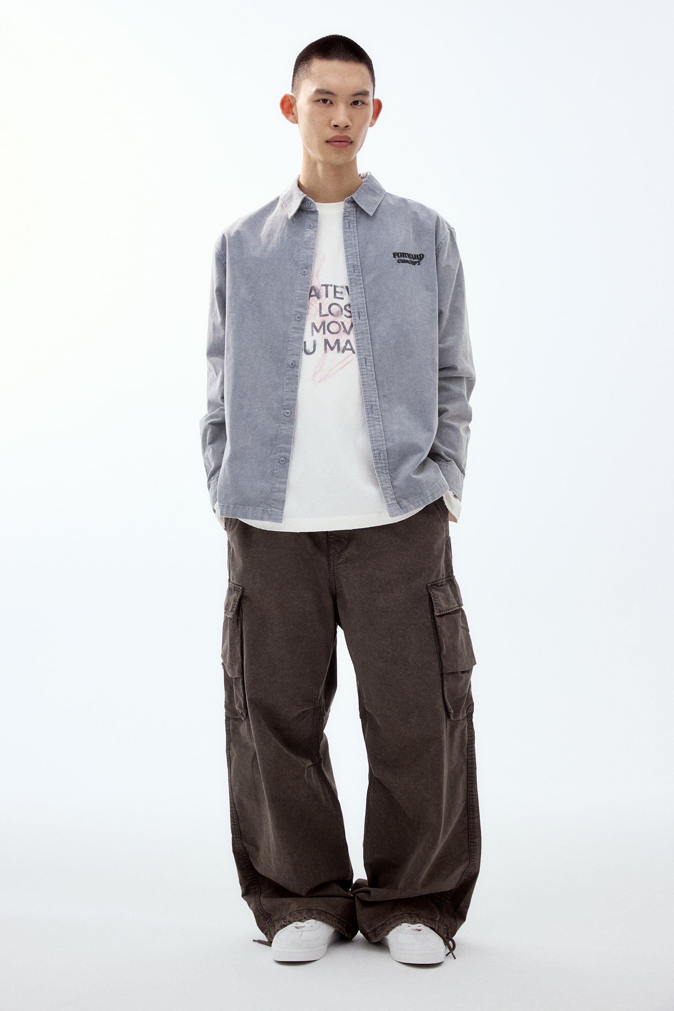 이미지 확대 보기: 모델이 정면을 향해 서 있으며, 은은한 텍스처의 연회색 긴팔 셔츠를 흰색 그래픽 티셔츠 위에 풀어 입고 있습니다. 또한 넉넉한 와이드 실루엣의 짙은 갈색 카고 팬츠(큰 사이드 포켓 포함)와 흰색 레이스업 스니커즈를 착용하고 있습니다.