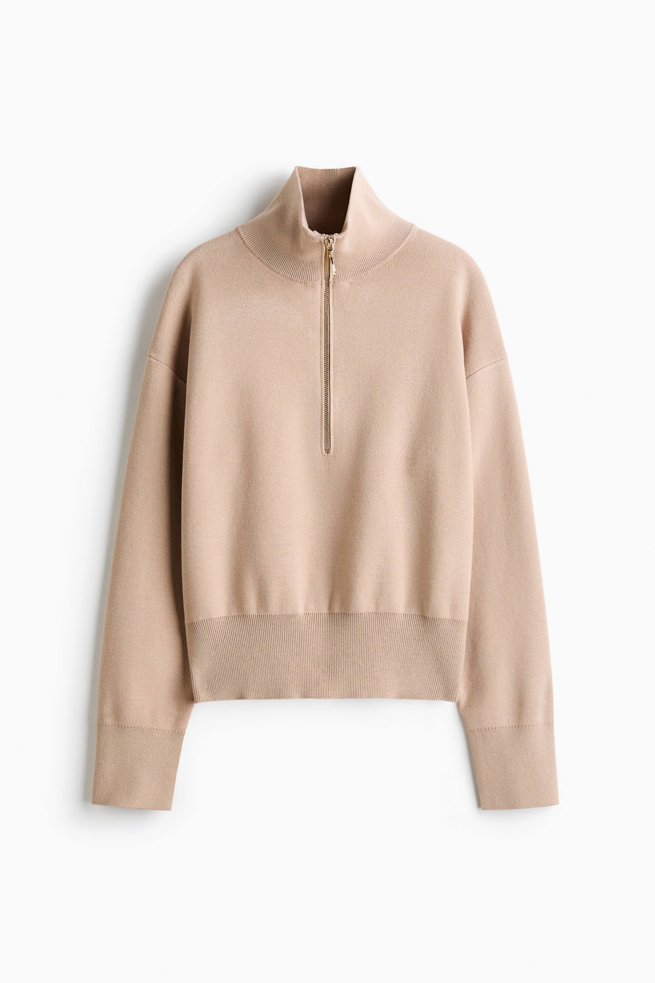 Visa större bild: En beige finstickad tröja visas framifrån, med en hög ribbad krage och en halv dragkedja i guldfärg. Den är designad med sänkta axlar, långa ärmar med breda ribbade muddar och en matchande bred ribbad nederkant.