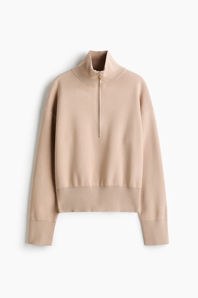 A beige fine-knit jumper is shown from the front, featuring a high ribbed collar with a gold-tone half-zip fastening. It is designed with dropped shoulders, long sleeves with wide ribbed cuffs, and a matching wide ribbed hem. - 1