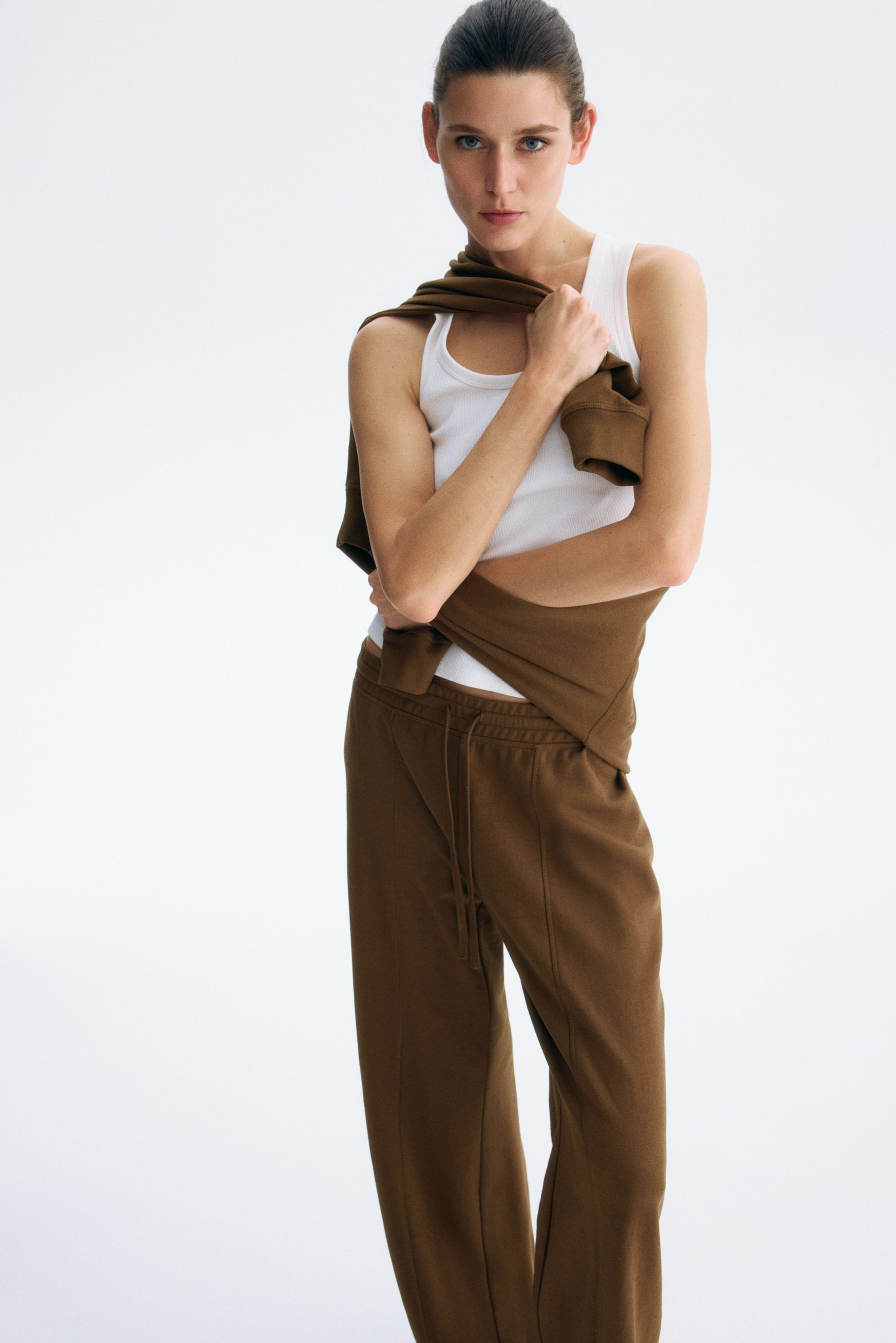 Xem ảnh lớn hơn: Quần jogger màu nâu được trình bày nghiêng nhẹ sang phải, nổi bật với cạp quần có dây rút và các đường may chạy dọc phía trước mỗi ống quần. Áo nỉ màu nâu được khoác lên vai và phần trên cơ thể, tay áo vắt ngang phía trước, trong khi áo ba lỗ màu trắng có gân được mặc bên dưới.