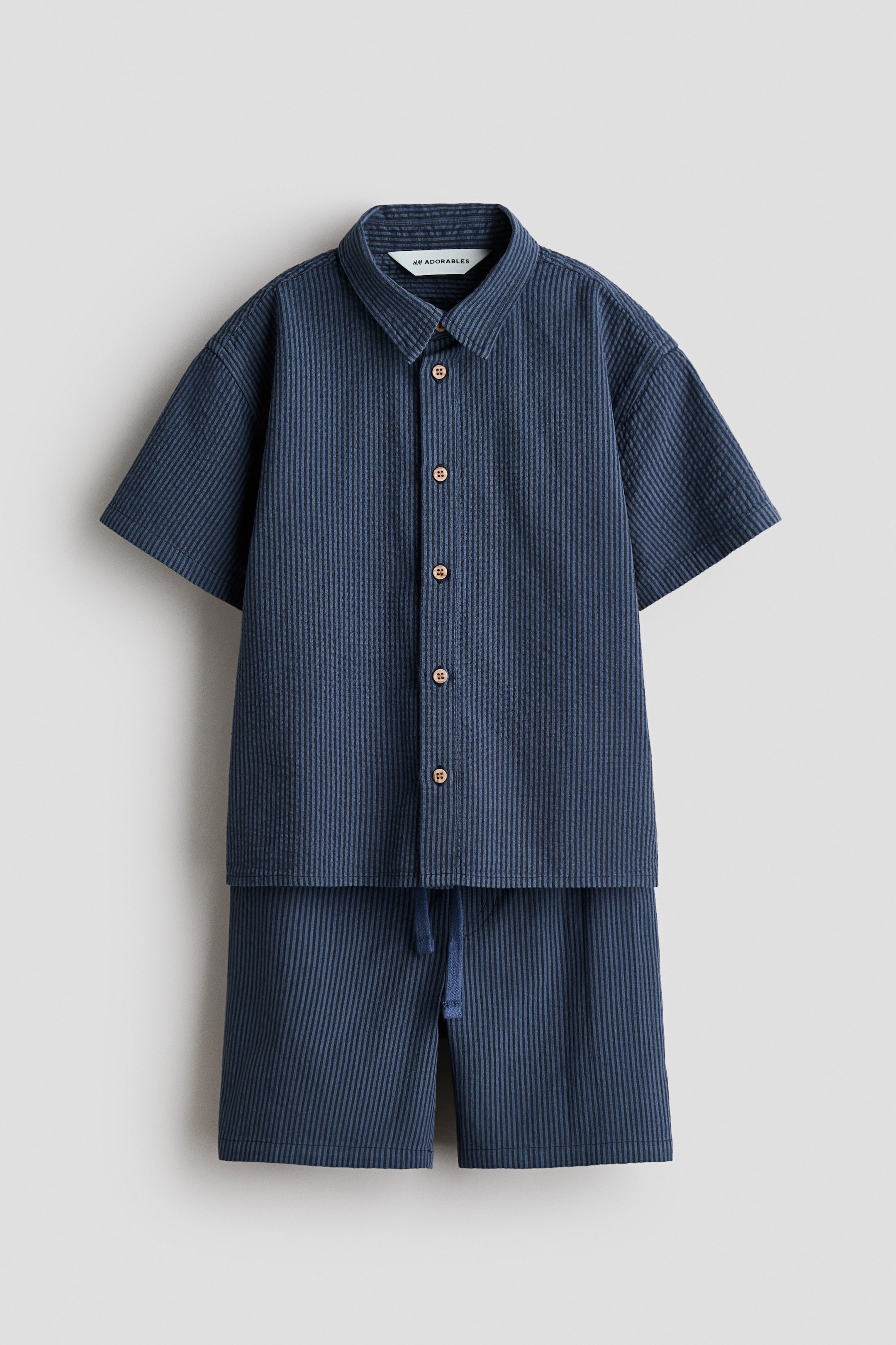 Agrandir l'image: Un ensemble en seersucker bleu foncé est présenté de face, comprenant une chemise à manches courtes avec un col rabattu, des boutons marron clair sur le devant et des manches tombantes, accompagnée d'un short assorti avec un cordon de serrage visible à la taille.