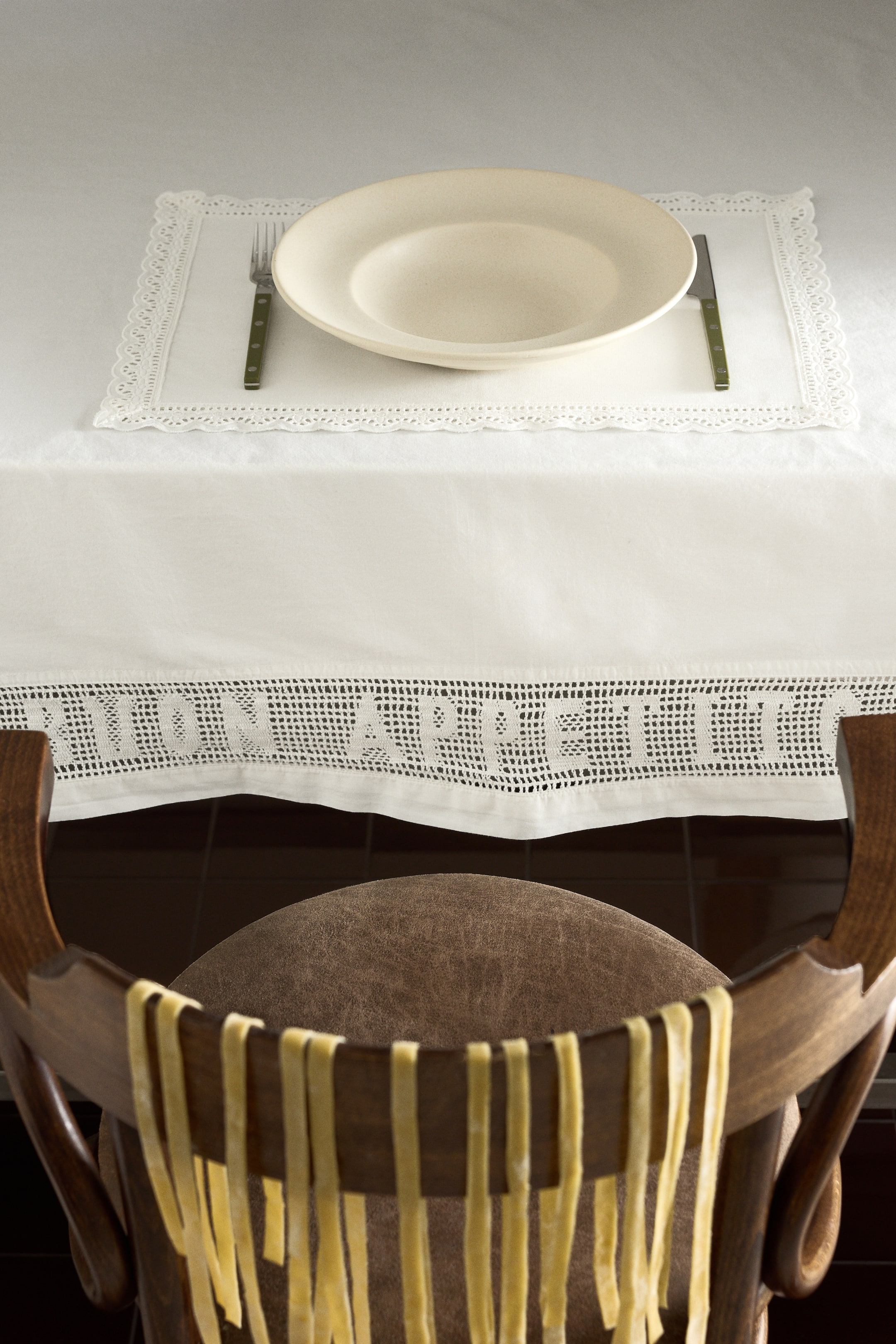 View larger image: White table linens adorn a table, featuring a rectangular placemat with a delicate lace border and a tablecloth with an openwork crochet lace band and text along its bottom edge. A cream-colored shallow serving plate is centered on the placemat, with a fork and knife resting beside it.