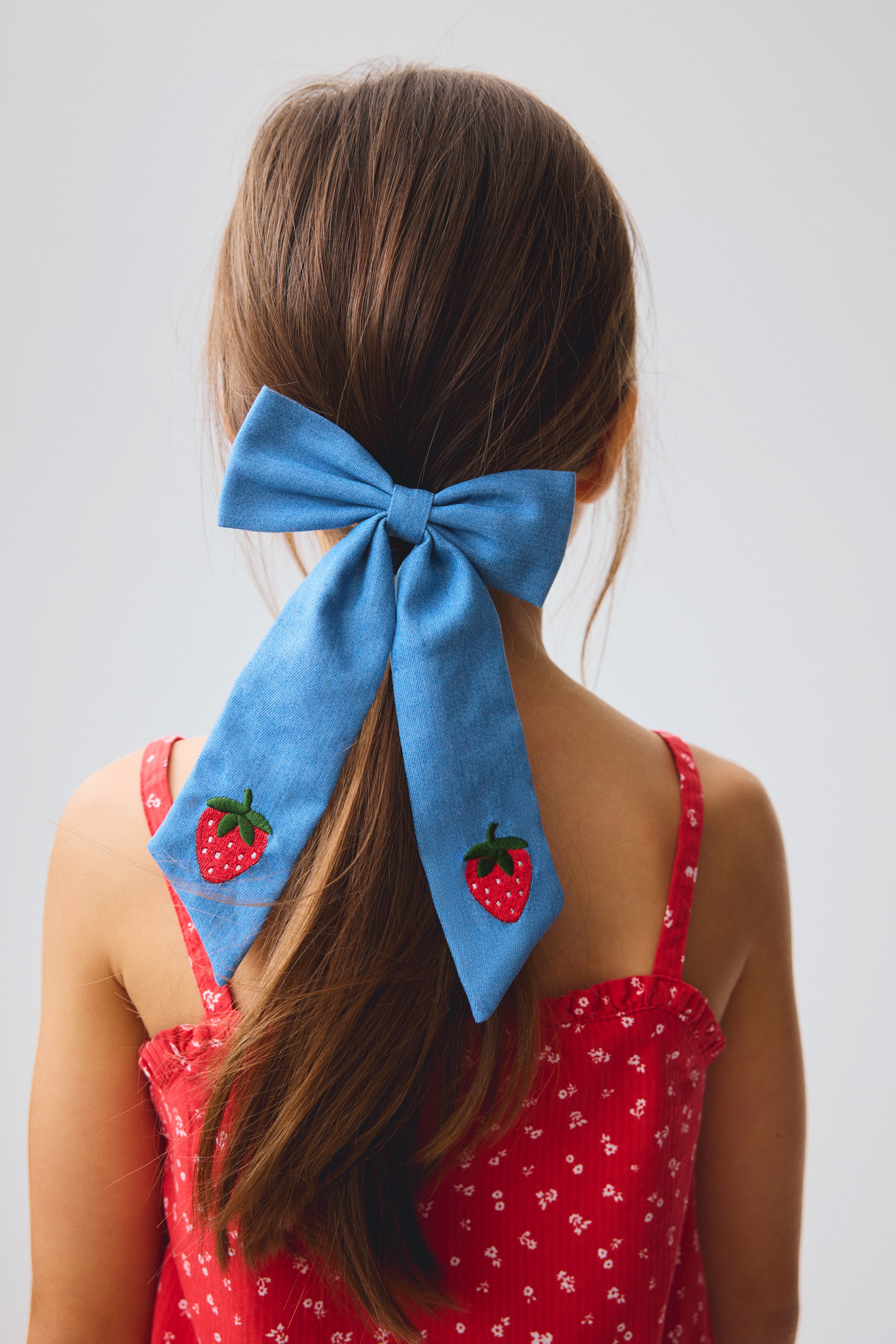 Zobacz większy obraz: Jasnoniebieska kokarda do włosów, przypominająca denim, jest zawiązana z tyłu głowy, utrzymując włosy na miejscu. Kokarda ma dwa długie ogony, z których każdy ozdobiony jest haftem czerwonej truskawki z zieloną łodyżką.
