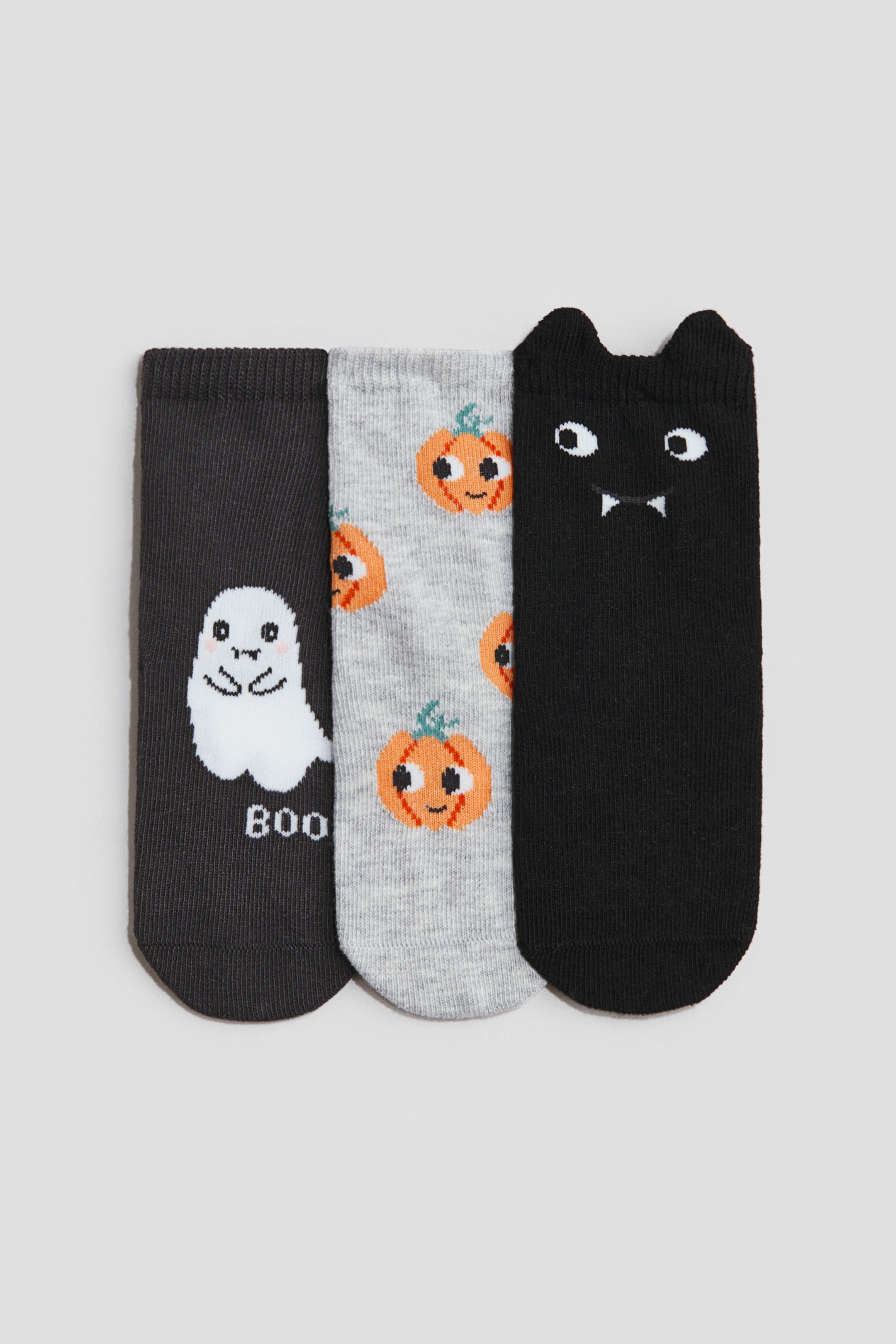 Voir une image plus grande: Les trois chaussettes à la cheville sont disposées à plat, chacune arborant des motifs individuels sur le thème d'Halloween. La chaussette gris foncé affiche un fantôme blanc de dessin animé avec le texte « BOO », la chaussette gris clair présente un motif intégral de citrouilles orange, et la chaussette noire met en avant un visage de chat noir avec des yeux blancs et des crocs, avec sa bordure formée en oreilles pointues.