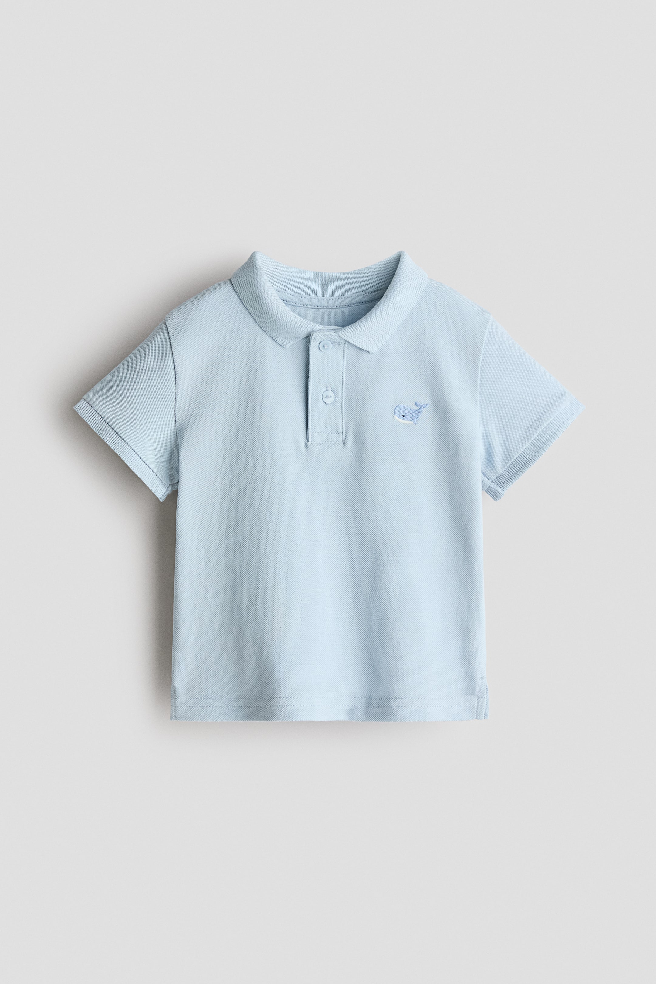 View larger image: A light-blue polo shirt is shown facing front, featuring a folded collar, a two-button placket, short sleeves with ribbed cuffs, and an embroidered whale motif on the left chest.