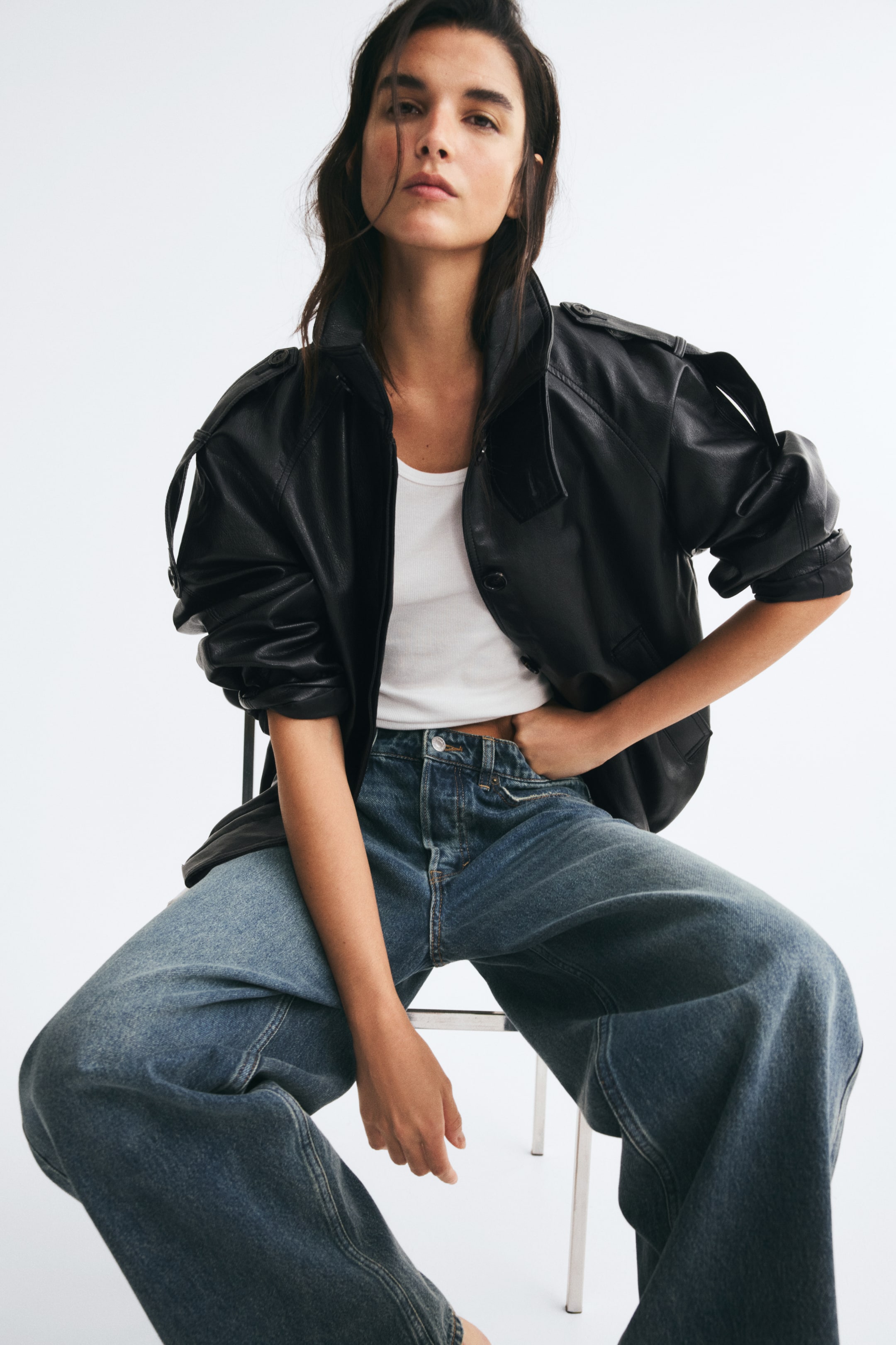 View larger image: A black oversized jacket in coated fabric is worn open by the model, layered over a white tank top and blue jeans. The jacket features a collar, buttoned shoulder tabs, and long sleeves pushed up to the elbows.