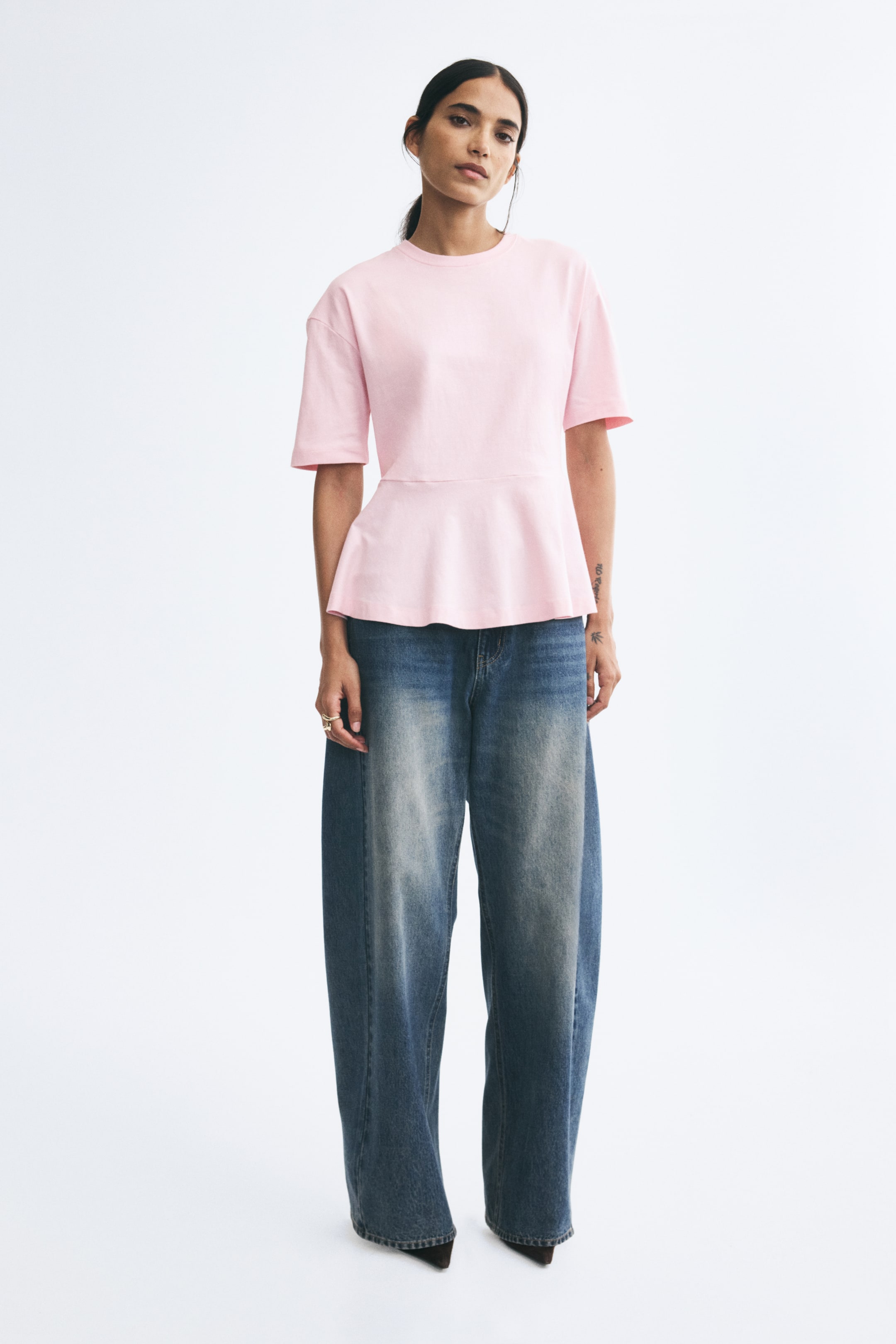 View larger image: The model is wearing a light pink cotton jersey top featuring a round neckline, dropped shoulders, short sleeves, and a flared peplum hem. The top is paired with faded blue wide-leg jeans.