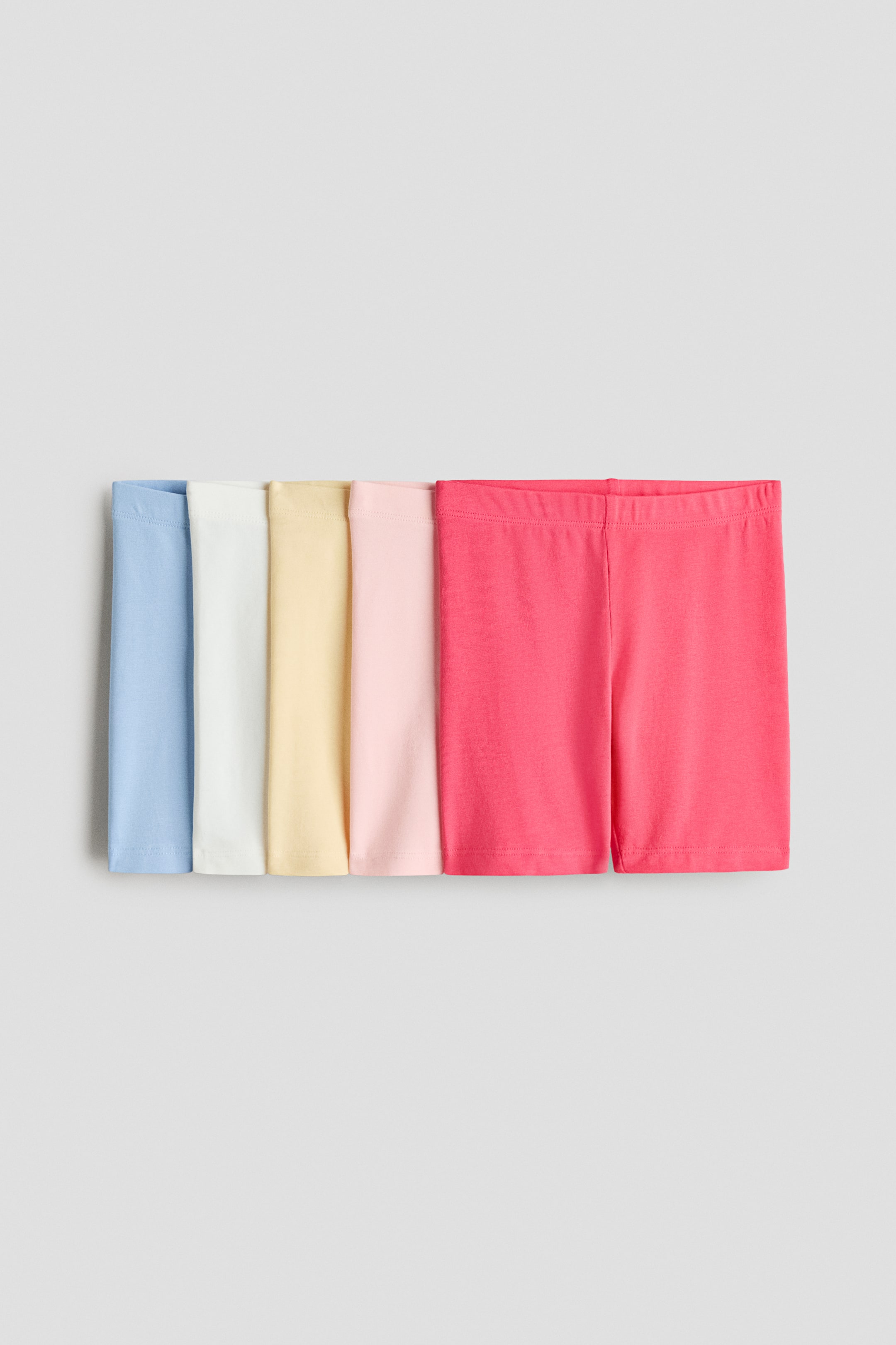 View larger image: A set of cycling shorts in soft cotton jersey with an elasticated waist is laid flat, arranged horizontally. They feature light blue, white, yellow, light pink, and bright pink colors.