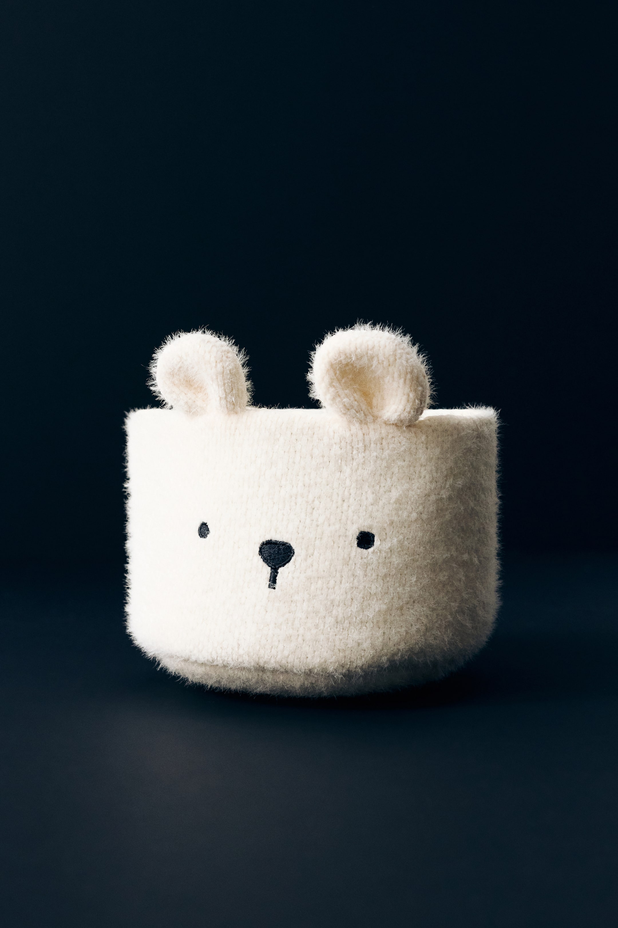 View larger image: A bear-design storage basket is shown from the front, featuring a cylindrical shape made of a soft, fuzzy off-white plush material. The basket has two rounded ears protruding from the top rim and embroidered black eyes and a nose on the front.