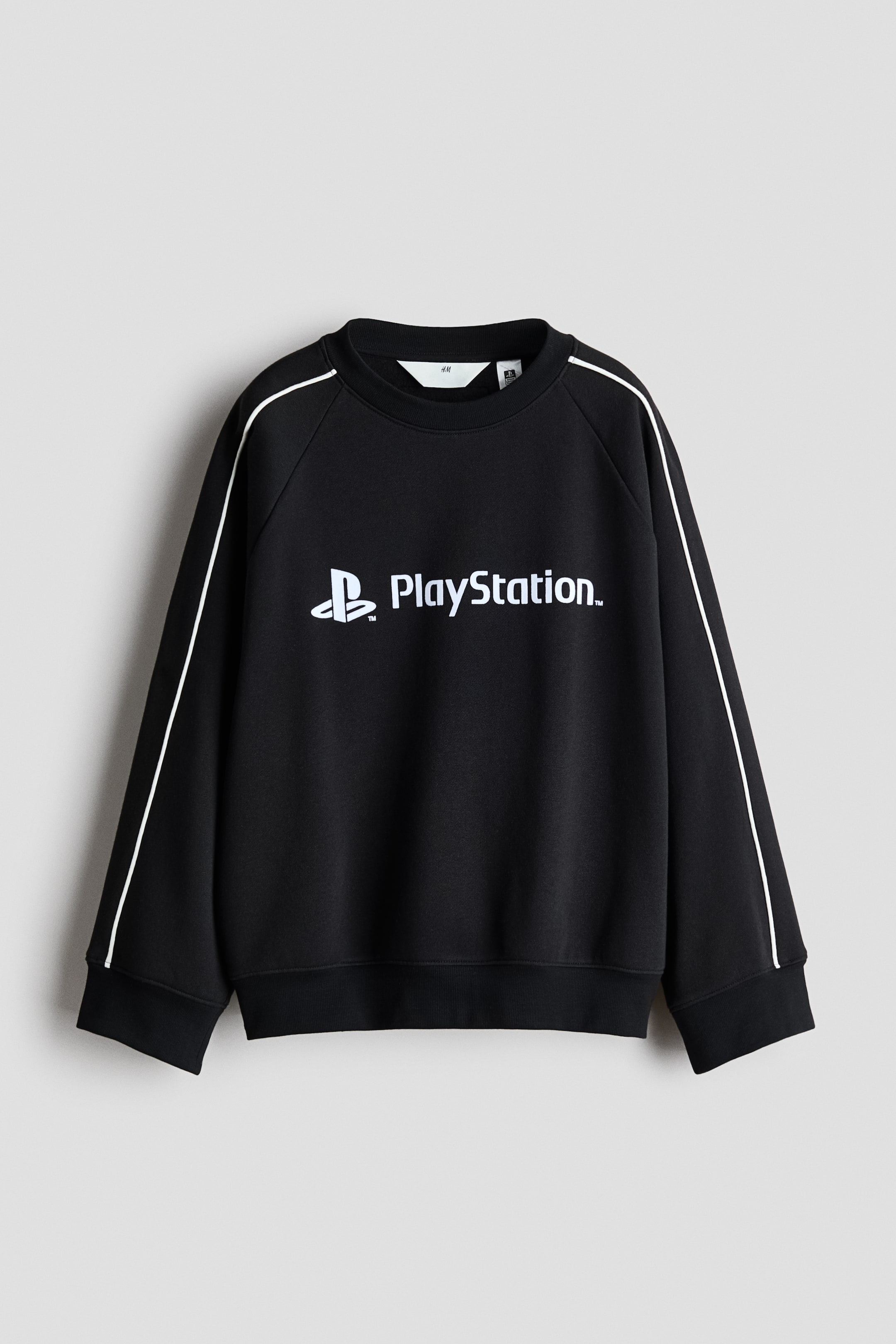 Visualizza immagine più grande: Una felpa nera con il logo PlayStation bianco e testo sul petto, con dettagli in piping bianco lungo le cuciture raglan delle maniche. È progettata con un girocollo, polsini a coste e un orlo a coste.
