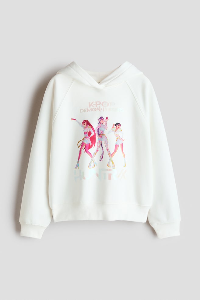 Ein oversized weiÃer Hoodie ist von vorne zu sehen und verfÃ¼gt Ã¼ber eine Kapuze mit Wickel-Design, lange RaglanÃ¤rmel sowie breite gerippte BÃ¼ndchen an Ãrmeln und Saum. Auf der Vorderseite befindet sich ein farbenfrohes, gedrucktes Motiv mit drei stilisierten animierten Figuren unter dem Schriftzug âK-POP DEMON HUNTERSâ. - 1