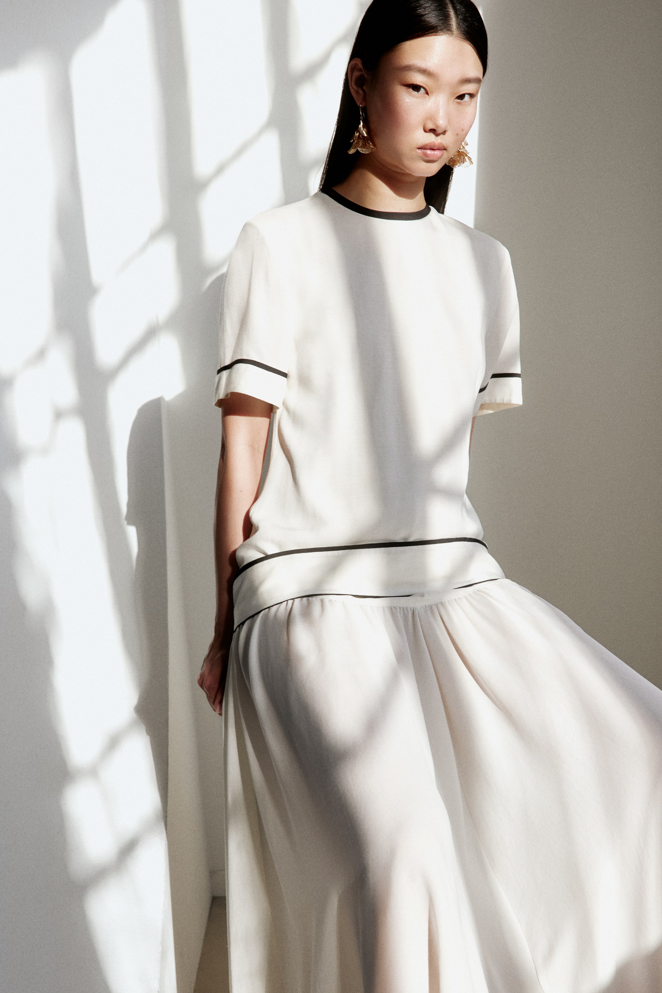 Agrandir l'image: Un haut à manches courtes de couleur blanc cassé est présenté, avec une bordure noire autour de son col rond, des poignets de manches et de l'ourlet. Une jupe longue et fluide assortie complète le haut, montrant une taille froncée avec une bordure noire sur son bord supérieur.