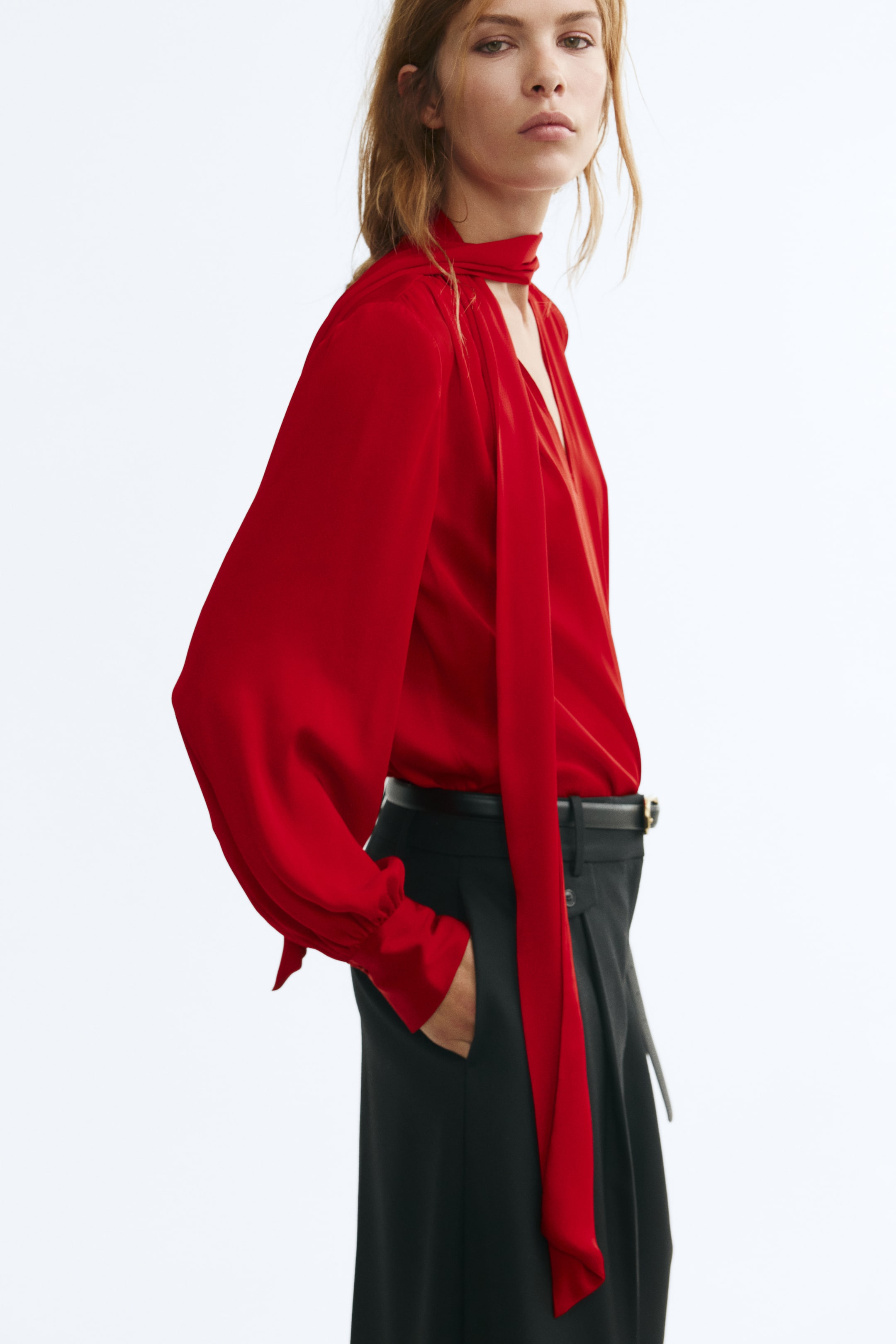 View larger image: A red woven blouse featuring a scarf collar with long hanging ties, a V-shaped front opening, and long balloon sleeves with button cuffs is worn by the model. The blouse is tucked into belted black trousers, and the model is standing with one hand placed in a pocket.