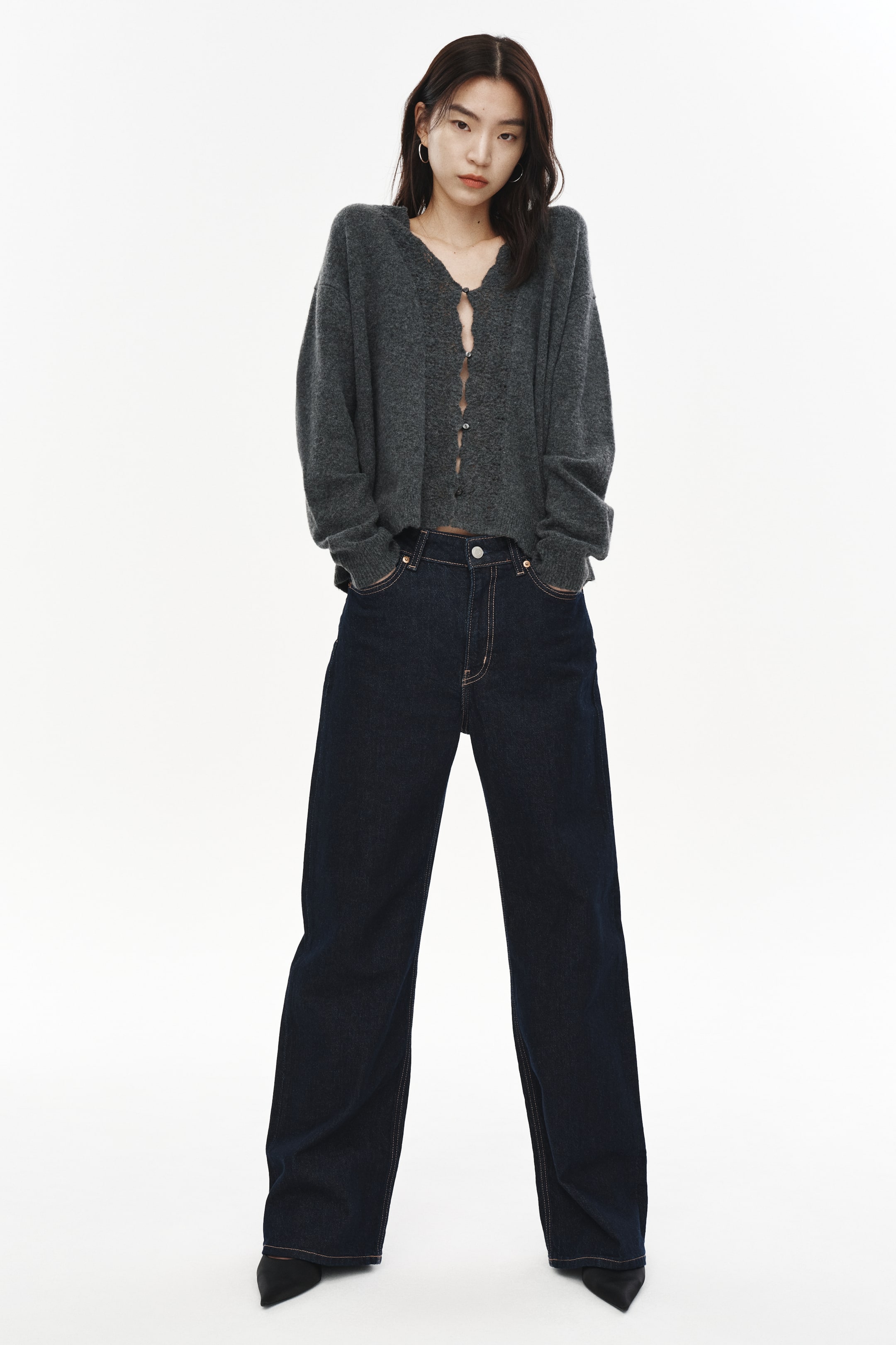 拡大画像を表示: モデルが着用しているダークグレーのカーディガンは、ソフトなニット素材で作られており、Vネックライン、前面にボタンがあり一部は留められています。ネックラインとボタンバンドには幅広のポワンテル編みトリムが施され、袖口と裾にはリブ編みが施されています。