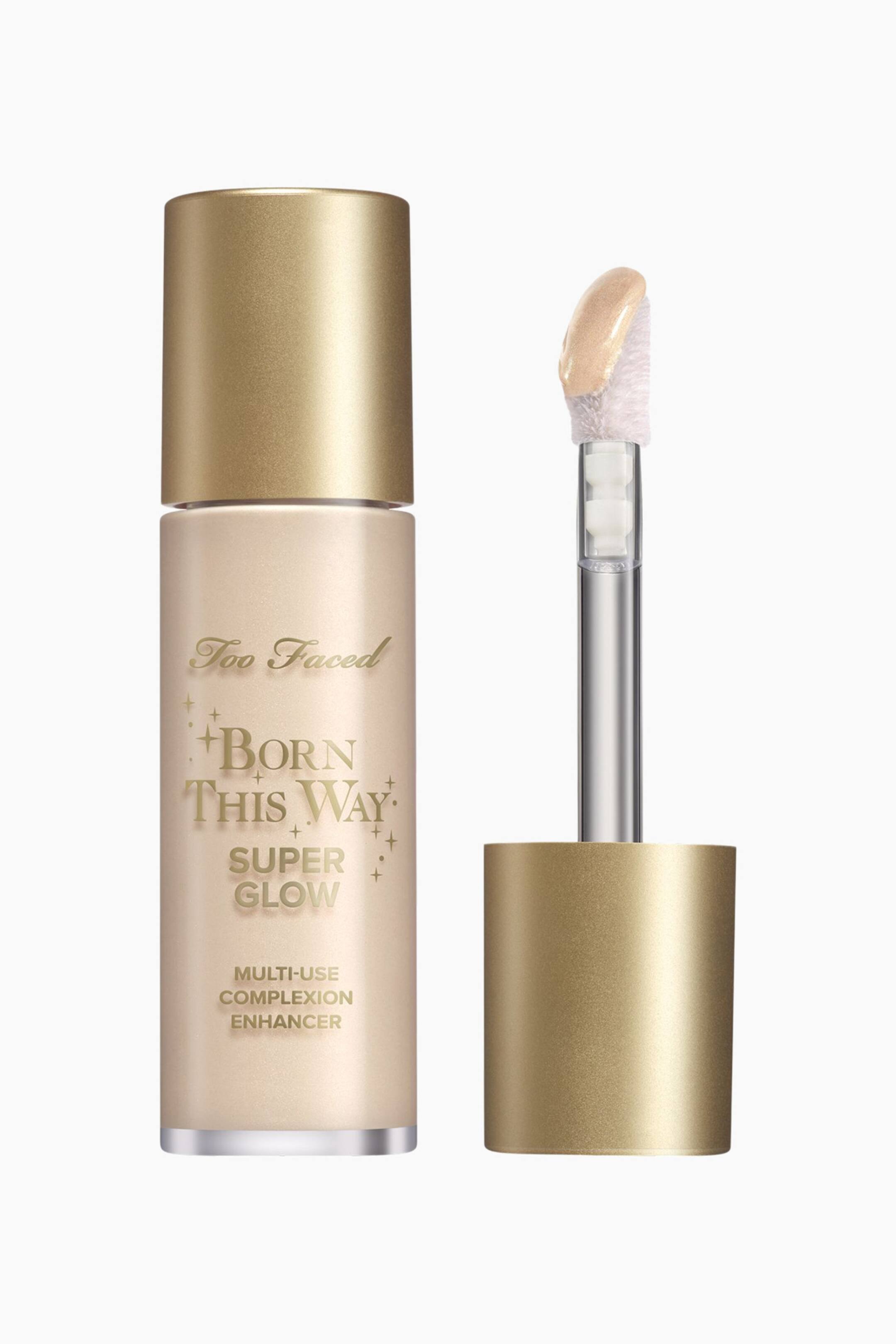Visa större bild: En ljusguldfärgad cylindrisk flaska av Too Faced Born This Way Super Glow Multi-Use Complexion Enhancer står upprätt, med guldfärgad text och en matchande guldkork. Bredvid står den separerade applikatorn också upprätt, med en guldgrund, en silverstav och en hjortfotstipp täckt i en skimrande ljusguldfärgad produkt.