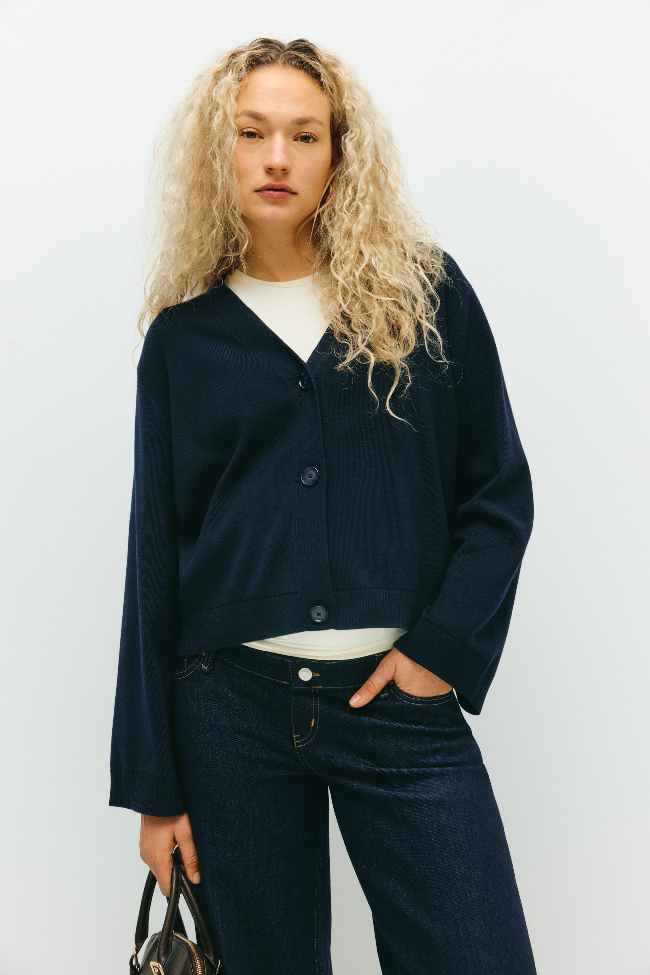 View larger image: A navy blue cardigan is being presented from the front, featuring a V-neckline, three dark buttons down the center, and long sleeves with ribbed cuffs. It is displaying a ribbed hem that ends above the waist, worn layered over a light-colored top.